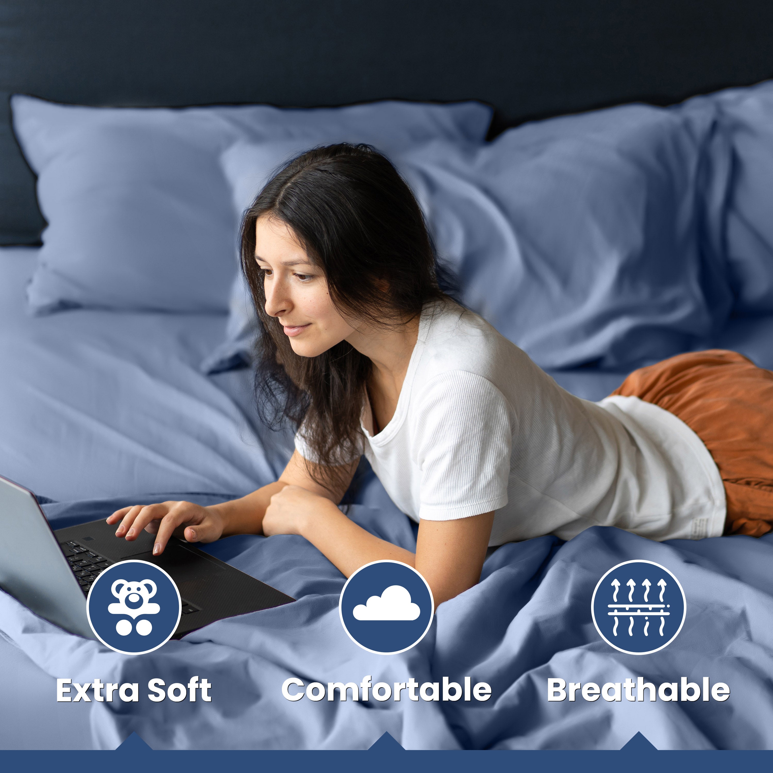 A woman using a laptop while lying on a bed with blue cotton sheets and pillows.