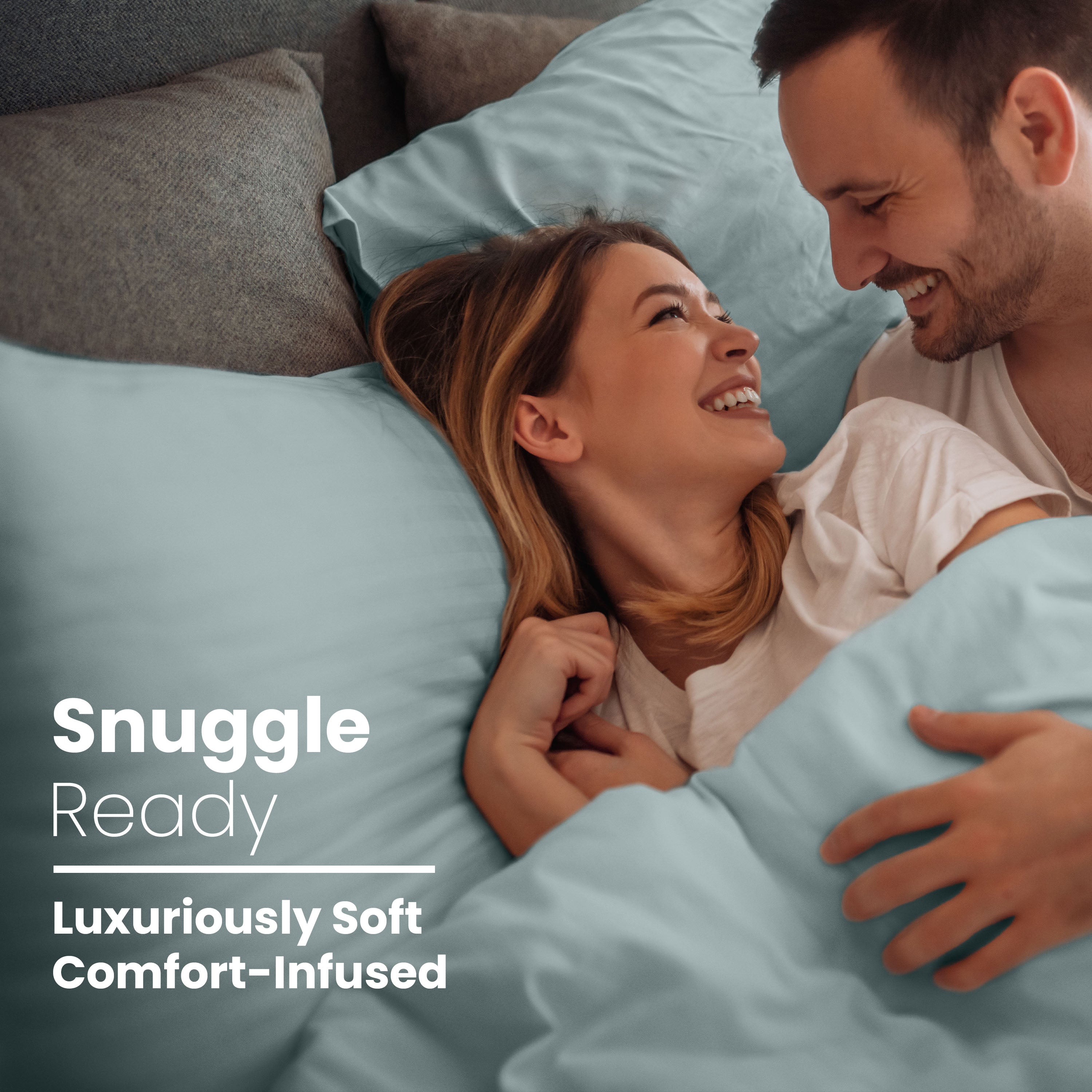 Couple smiling and cuddling under light blue sheets and pillows on a bed.