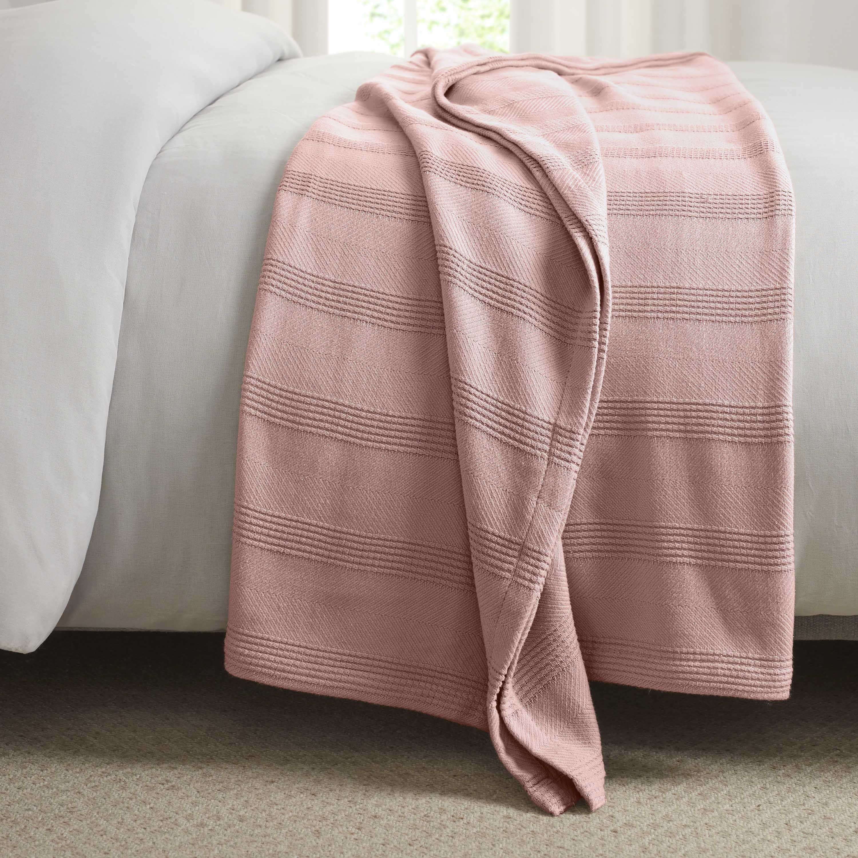 Cotton blanket in a soft pink color with textured stripes, draped over the edge of a bed.