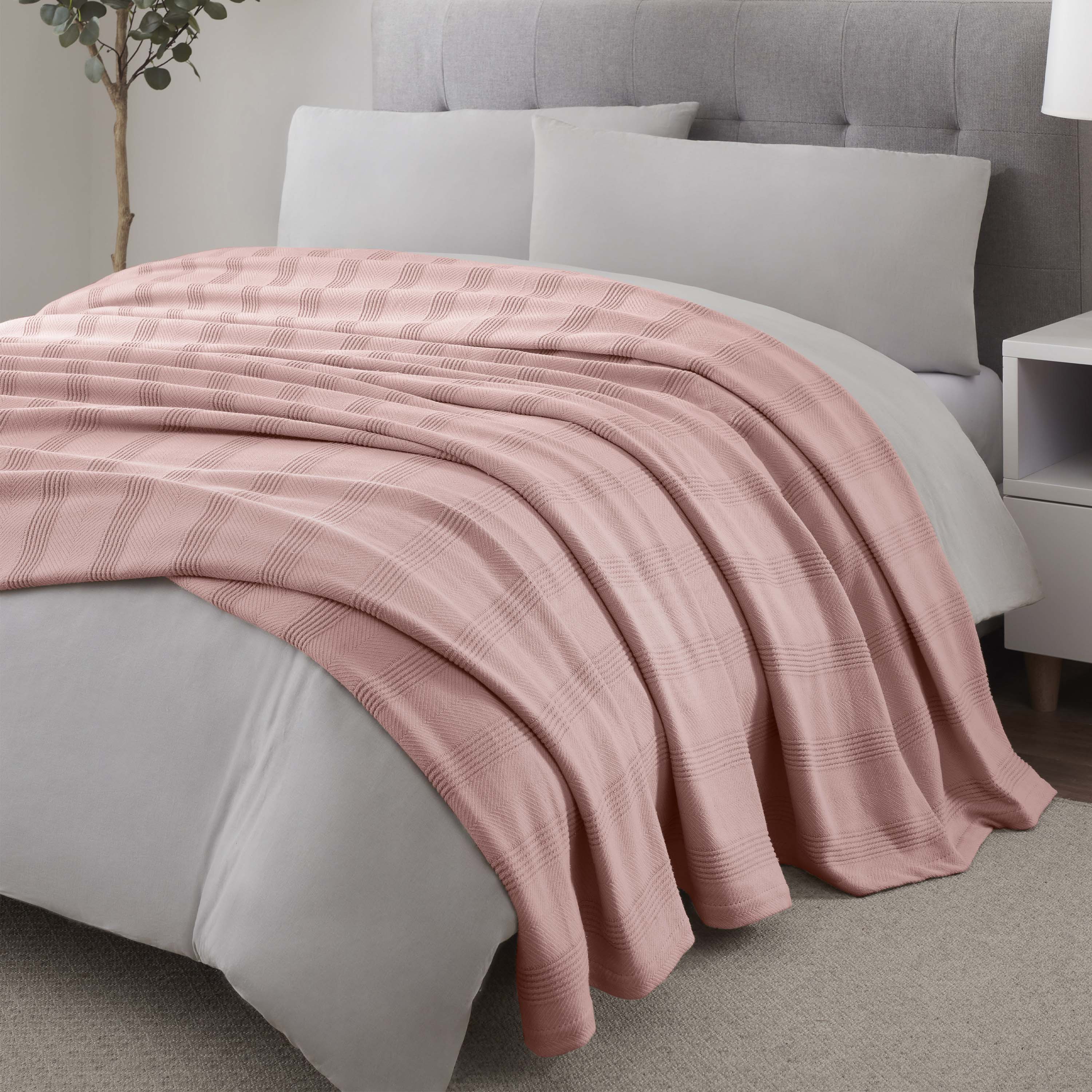 Light pink cotton blanket with a textured plaid pattern draped over a bed with gray bedding and pillows.