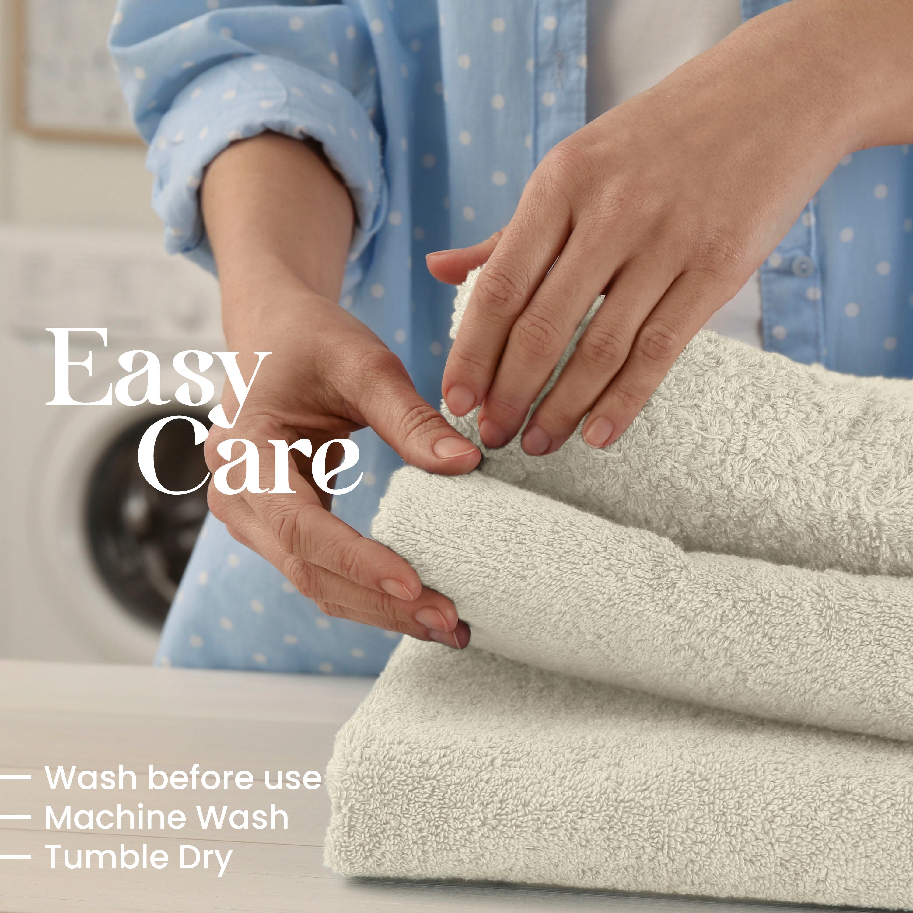 Hands folding soft, light-colored towels on a table, with a washing machine visible in the background.