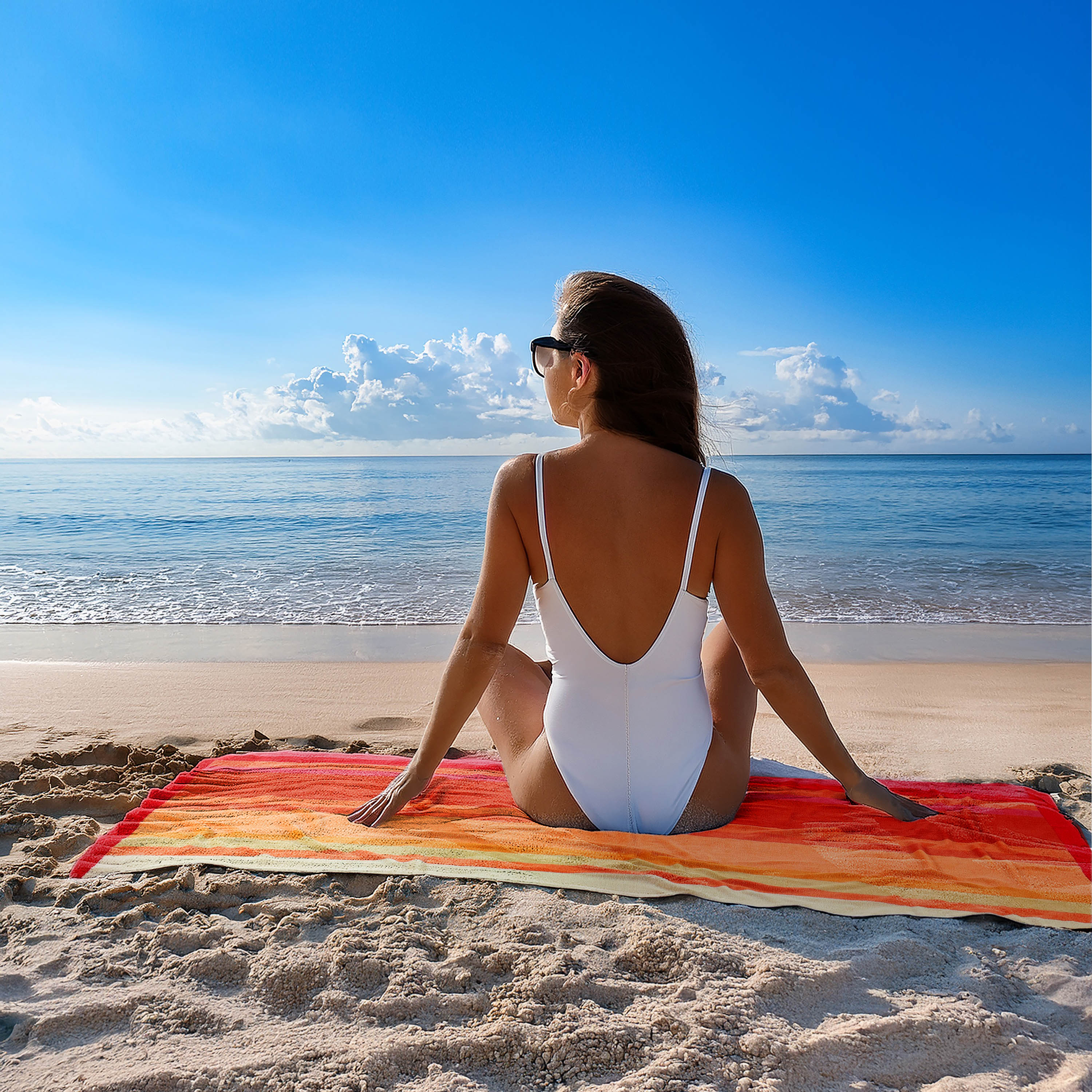 Woman in a white swimsuit sitting on a colorful towel at the beach with a clear blue sky.