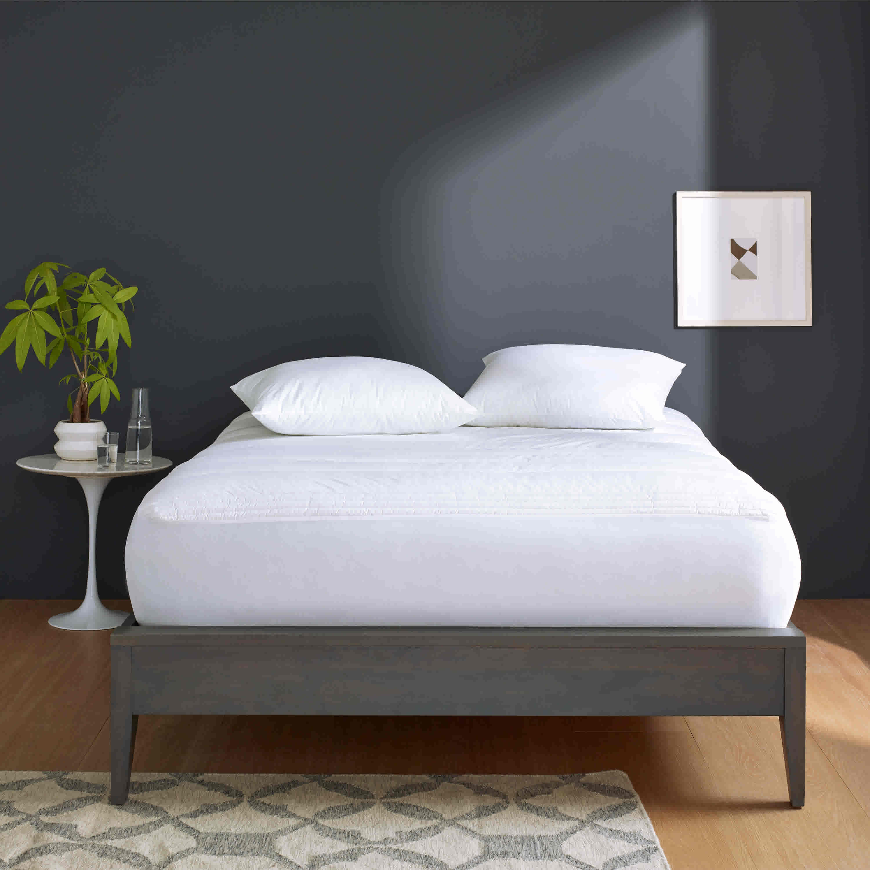 White mattress pad on a bed with two white pillows, a small side table, and a plant against a dark wall.