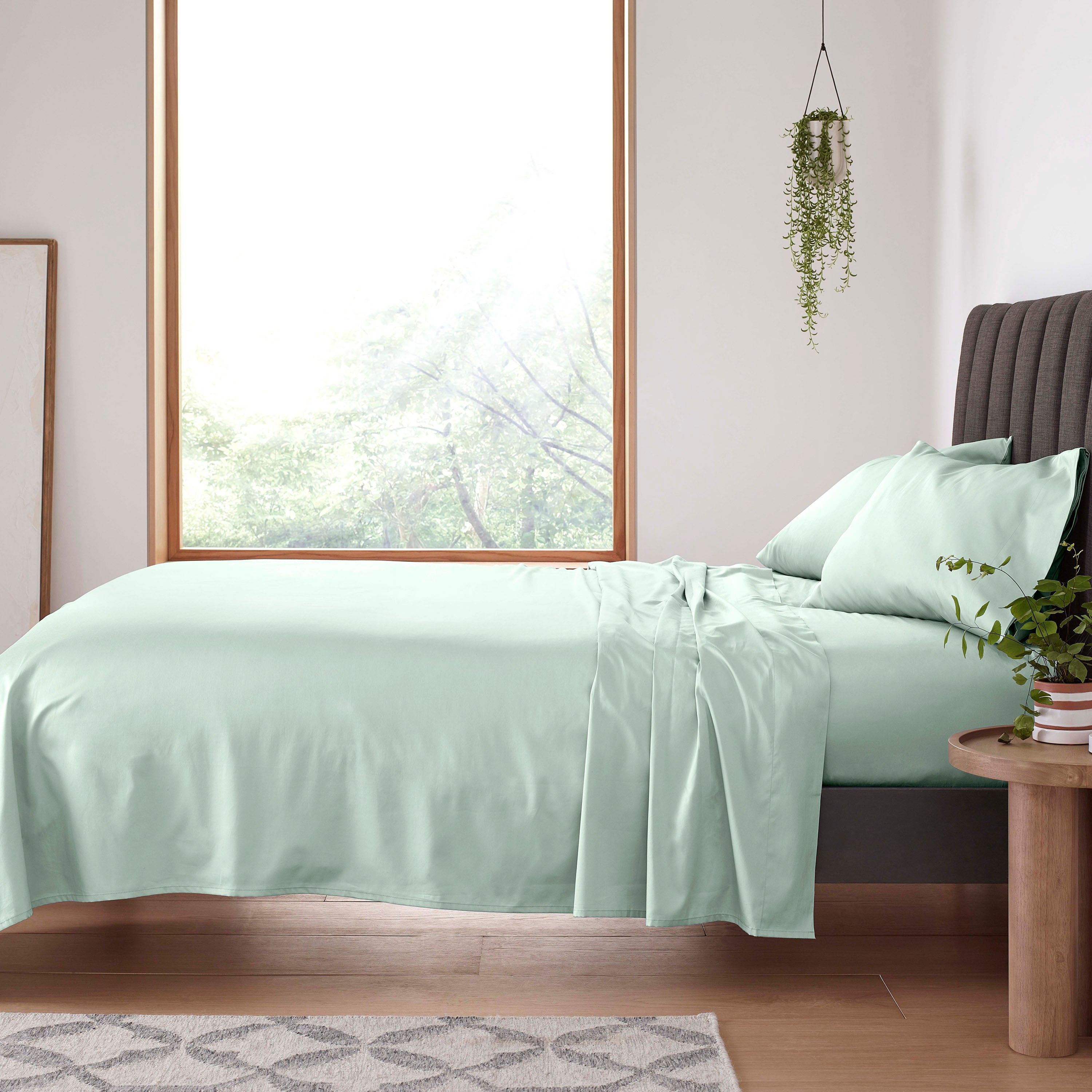 Light green sheet set laid on a bed with two matching pillows, beside a wooden side table and a window.