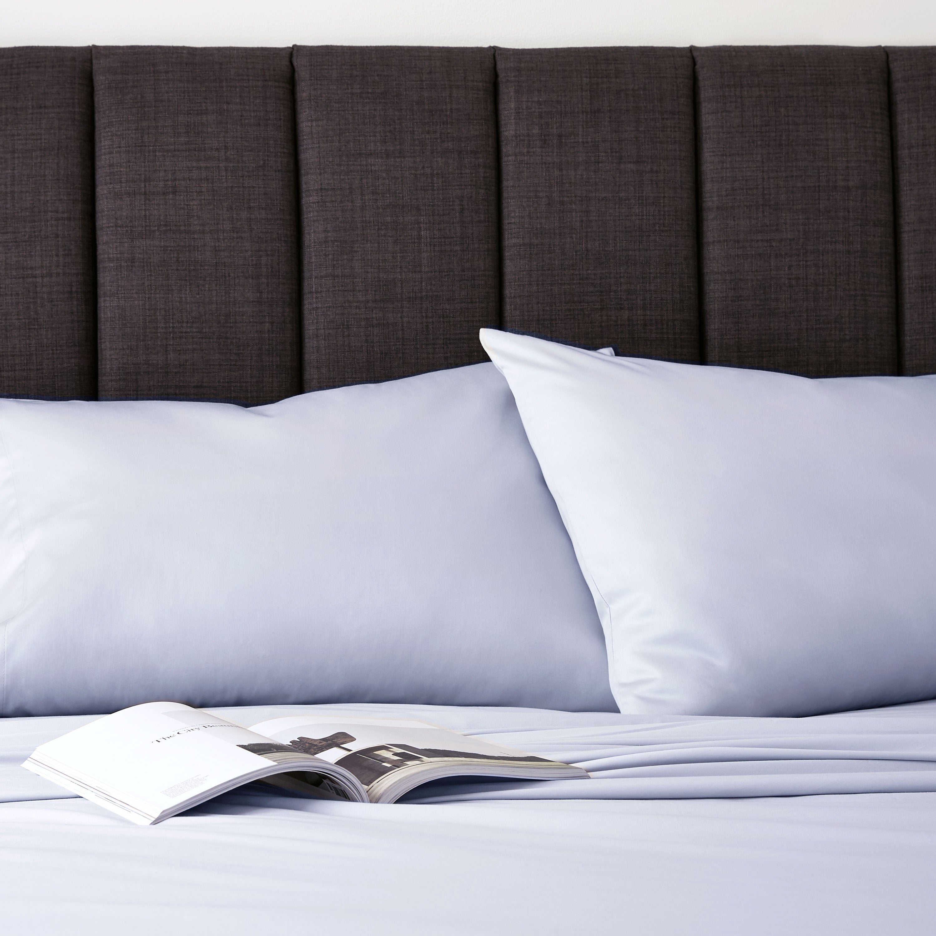 Light blue sheet set with two pillows on a bed, next to an open magazine and a dark upholstered headboard.