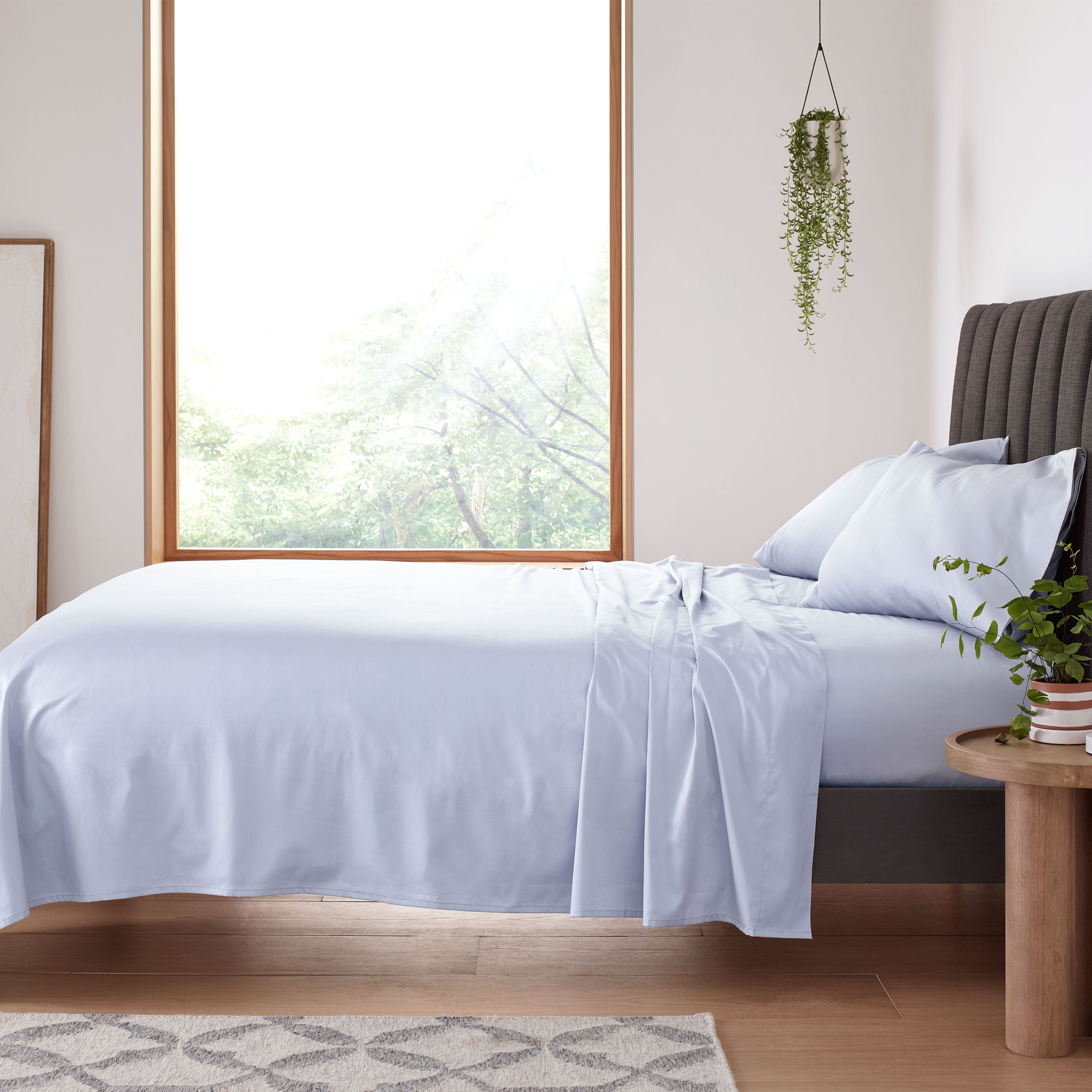 Light blue sheet set laid on a bed with two matching pillows, beside a wooden table and a window with greenery outside.