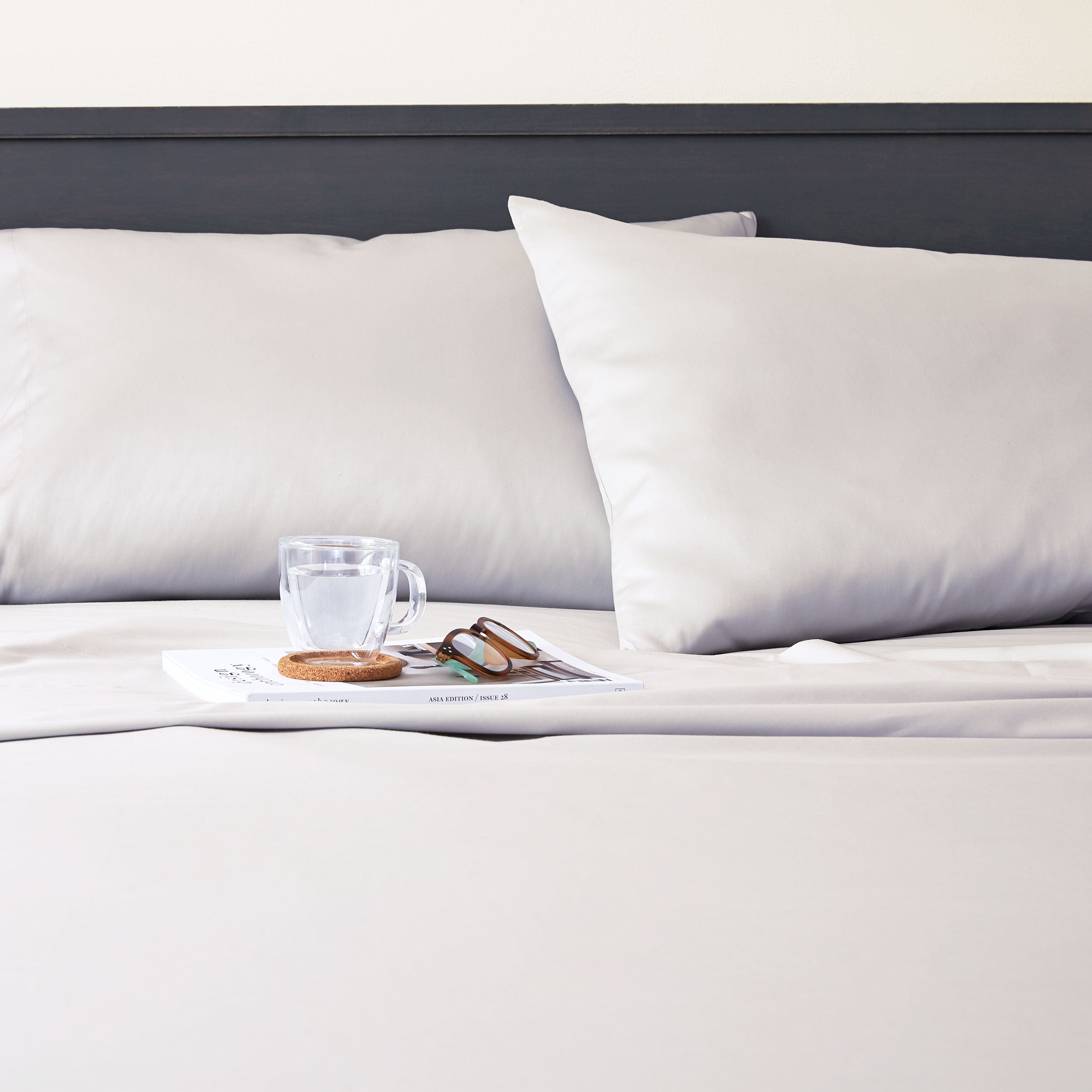 Light gray sheet set with two pillows, a glass of water, a coaster, and sunglasses on a magazine.