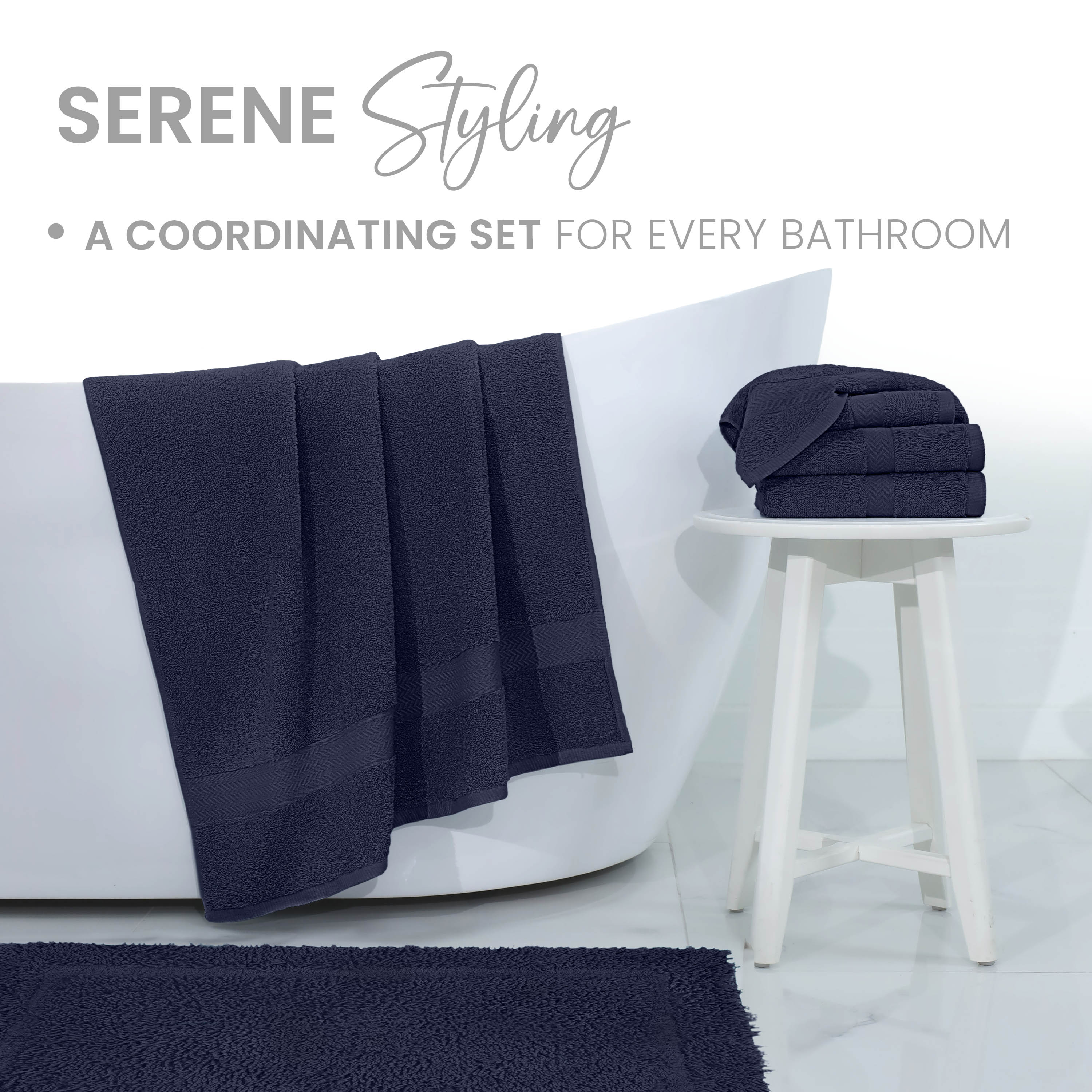 Navy bath rug displayed in a bathroom setting with towels hanging over a tub and stacked on a white stool.