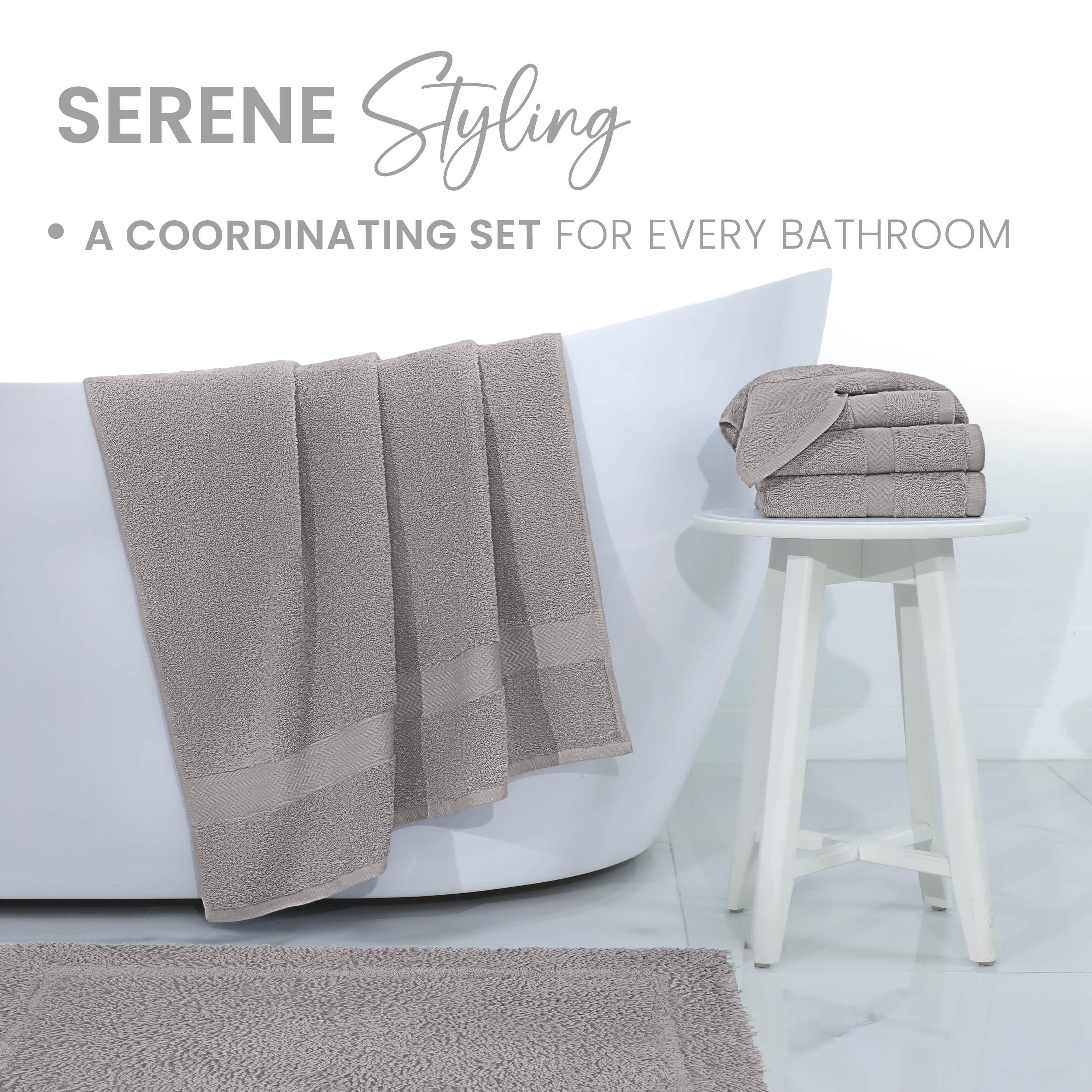 Gray bath rug laid on a tiled floor with matching towels hanging over a white bathtub and stacked on a small white stool.