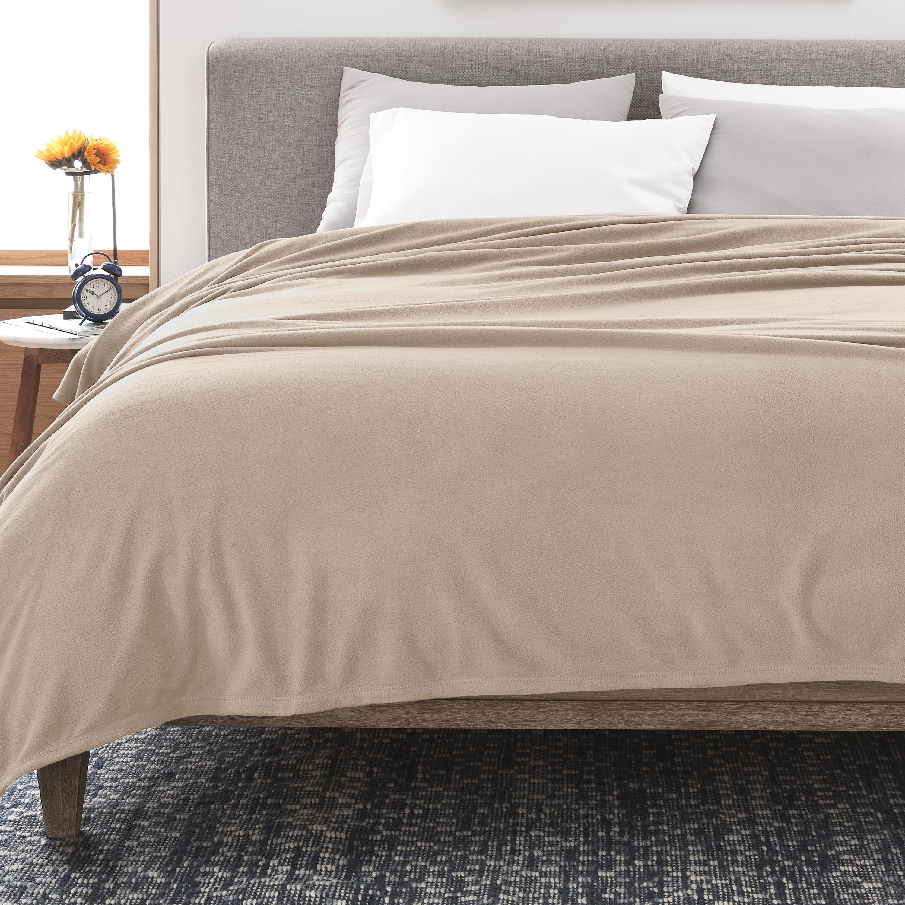 Soft beige fleece blanket draped over a bed with white and gray pillows, beside a nightstand with a clock and flower vase.