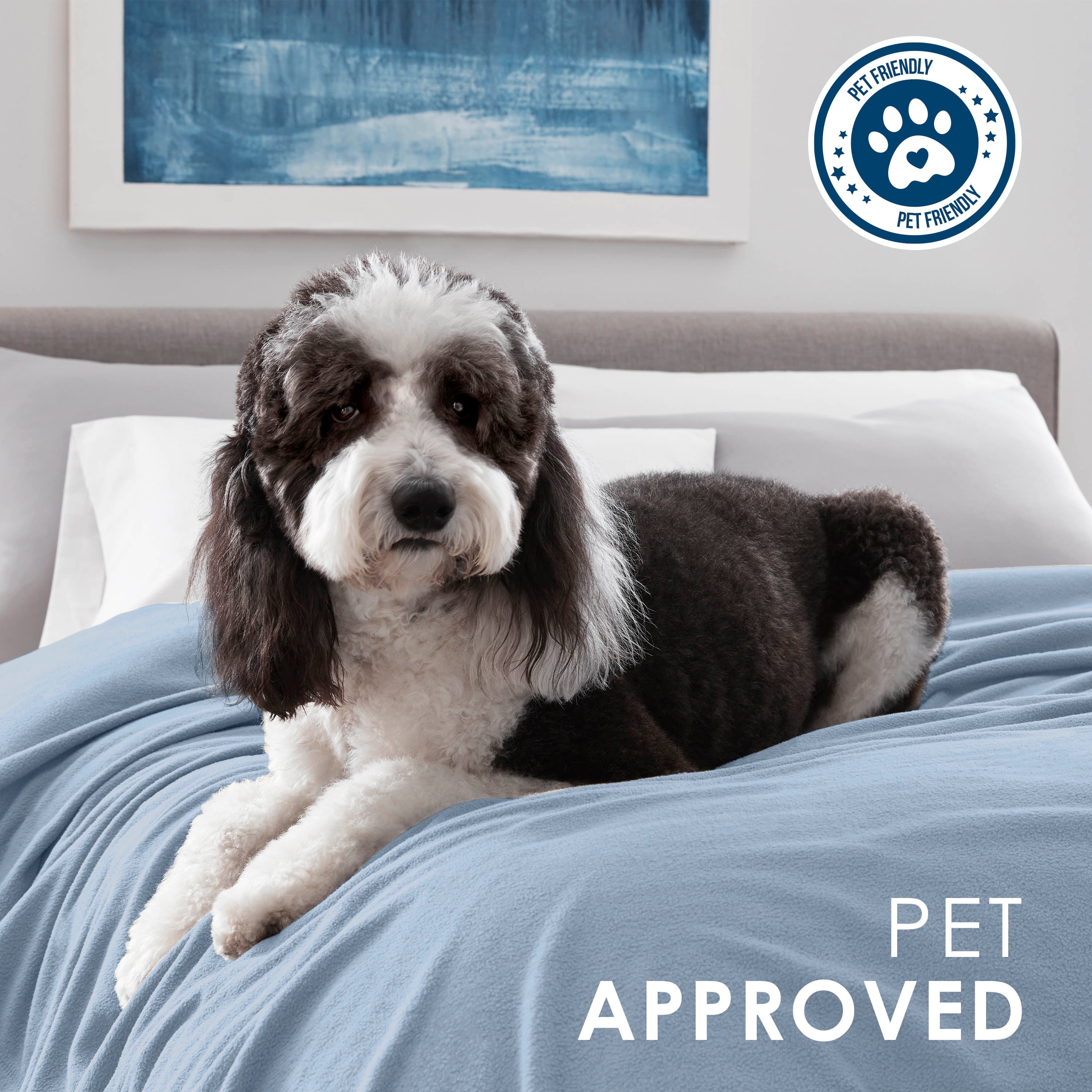 A black and white dog lies on a light blue fleece blanket on a bed with white pillows and a blue abstract painting in the bac