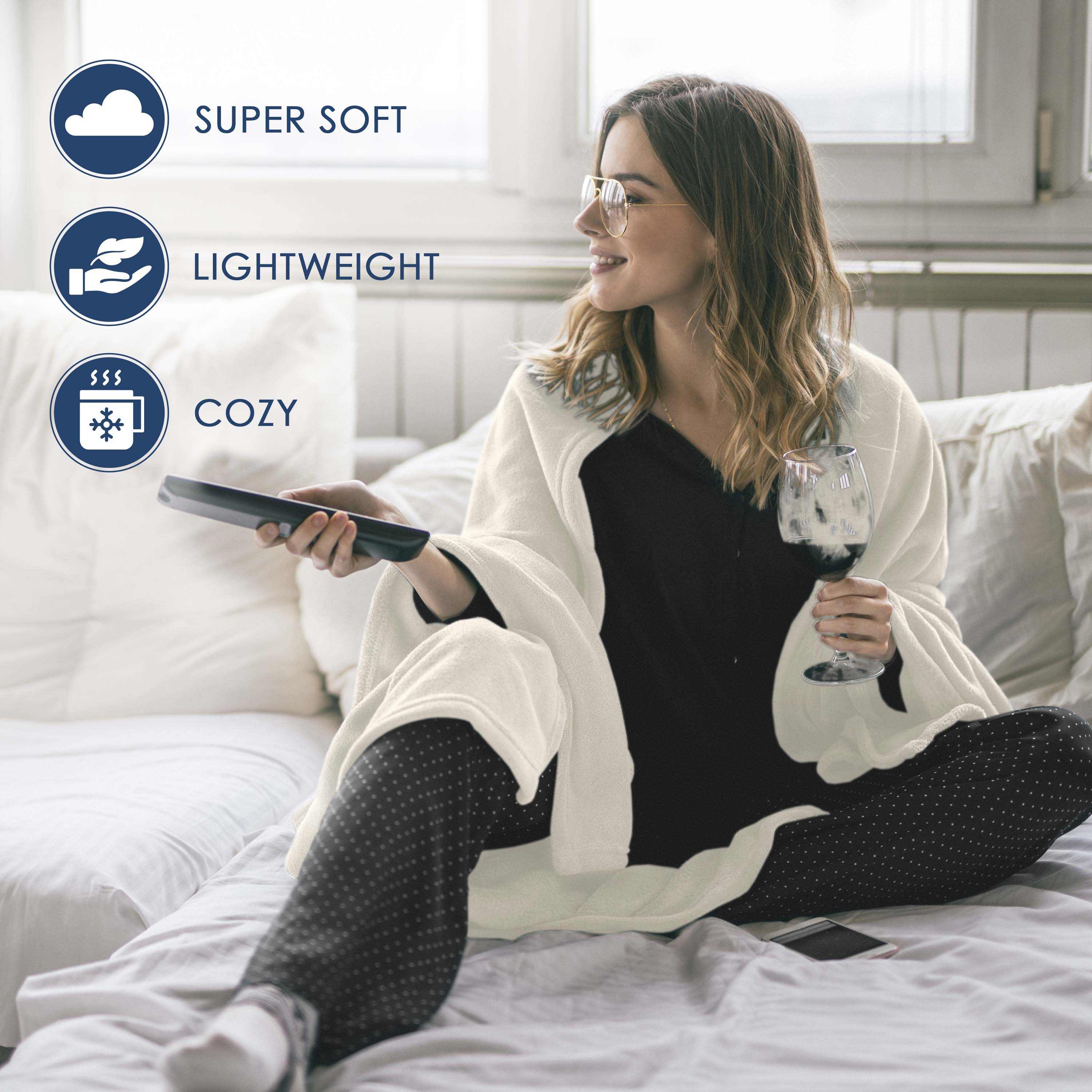 A woman sits on a bed wrapped in a white fleece blanket, holding a remote and a glass of wine.