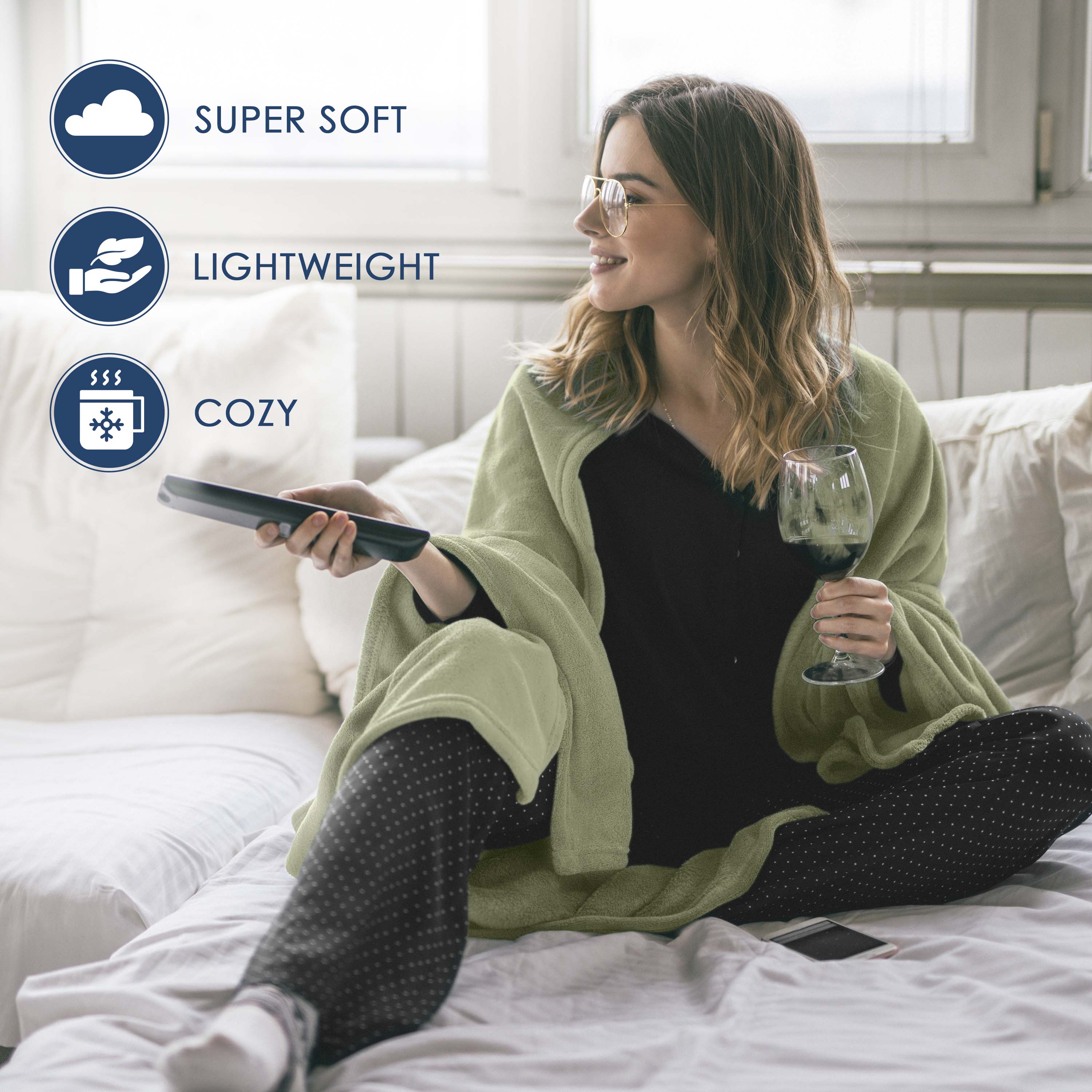 A woman sits on a bed wrapped in a light green fleece blanket, holding a remote and a glass of wine.