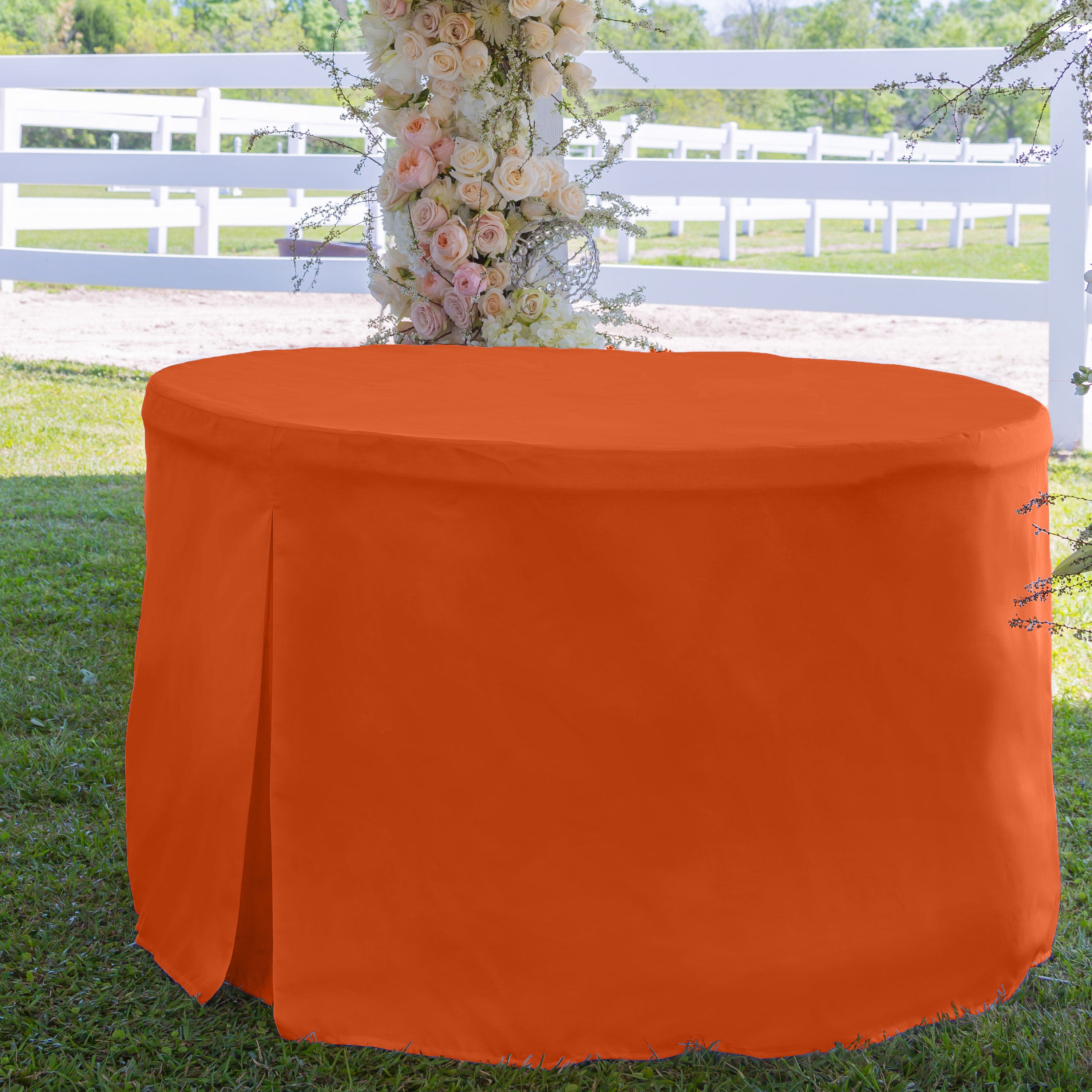 Round table cover in bright orange, fitting a 60-inch table, set on grass with a floral backdrop.