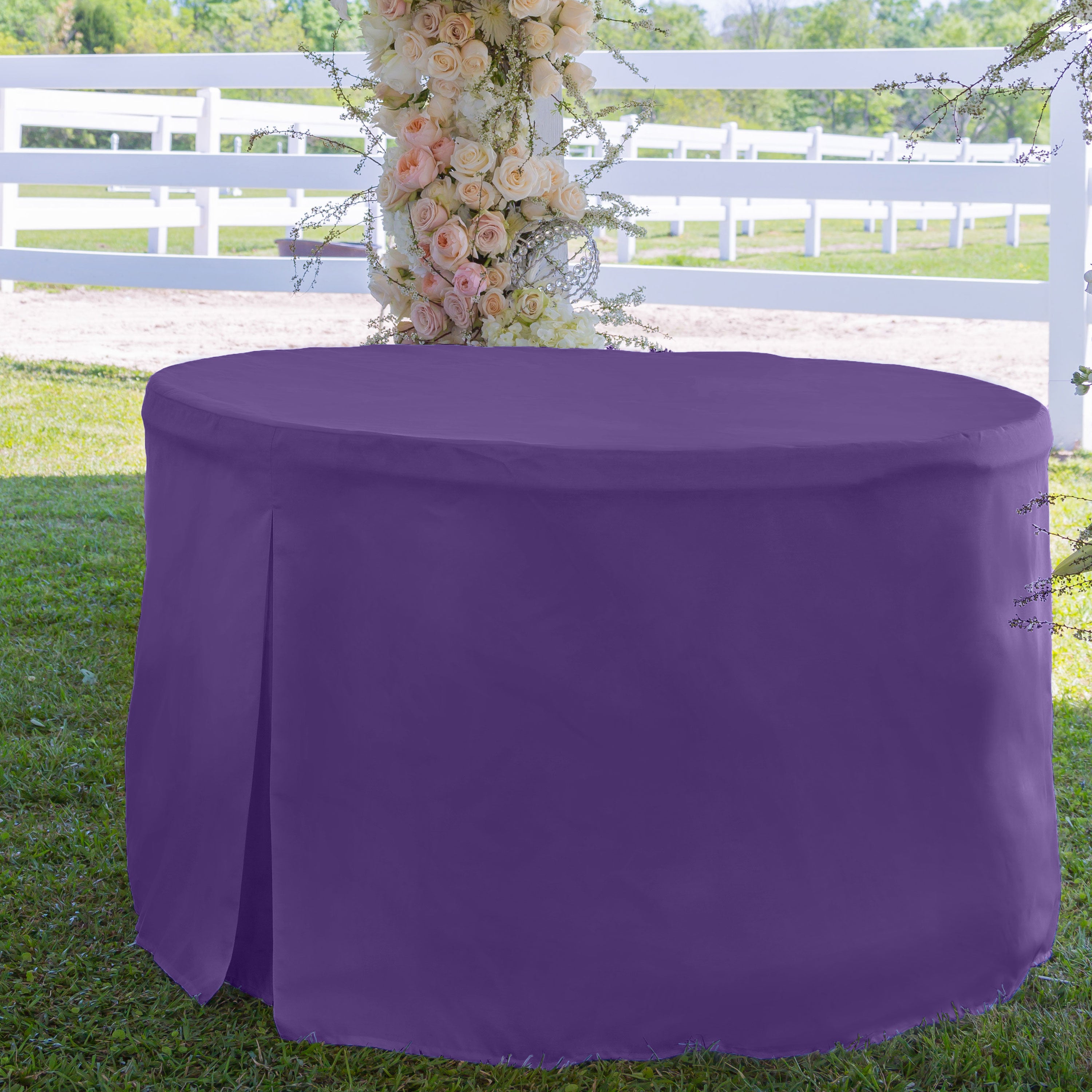 Round table covered with a solid purple tablecloth, set on grass with a floral backdrop.