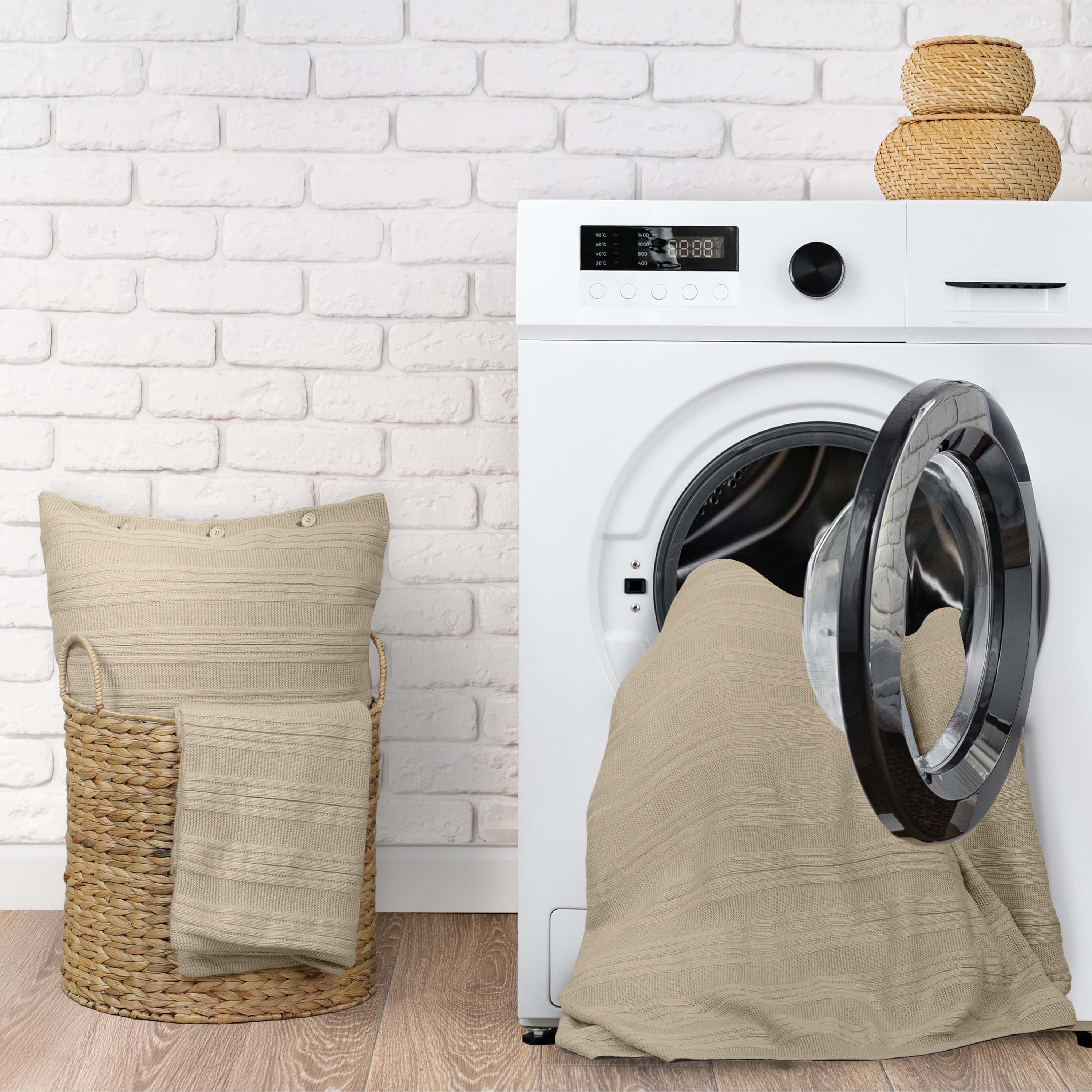 A beige blanket and pillow are placed near a washing machine against a white brick wall.