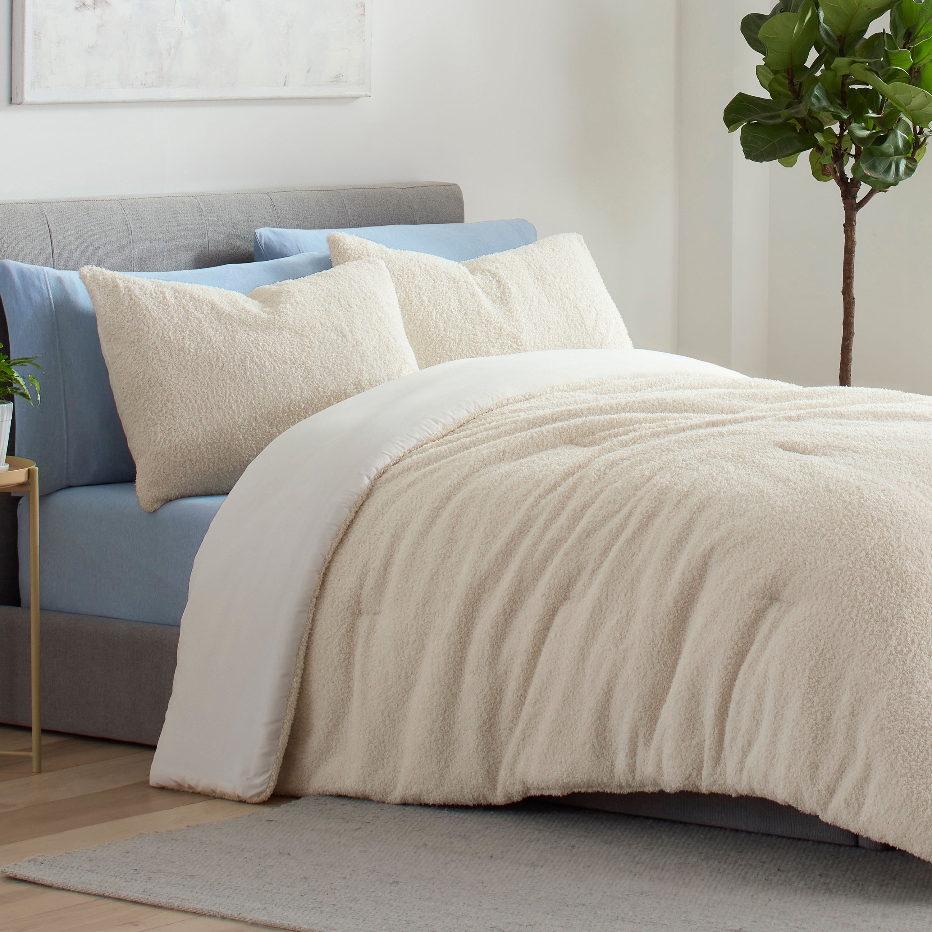 Cream-colored comforter set on a bed with blue and cream pillows, beside a small plant and a light-colored rug.