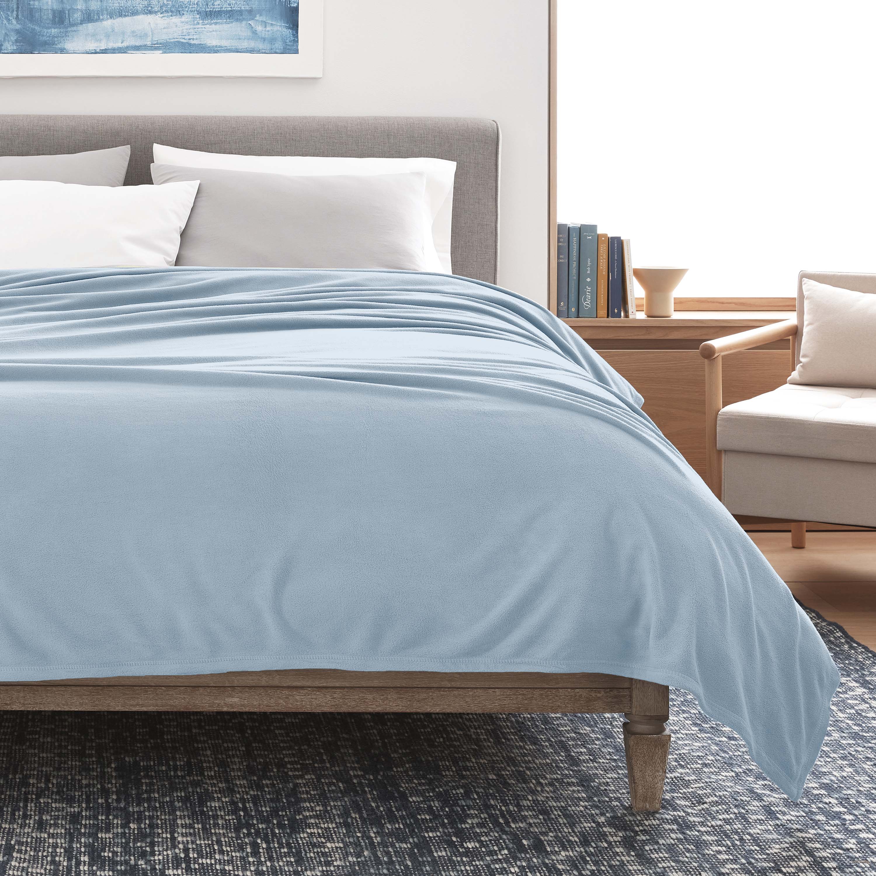 Light blue micro fleece blanket draped over a bed with gray headboard and white pillows, next to a wooden side table.