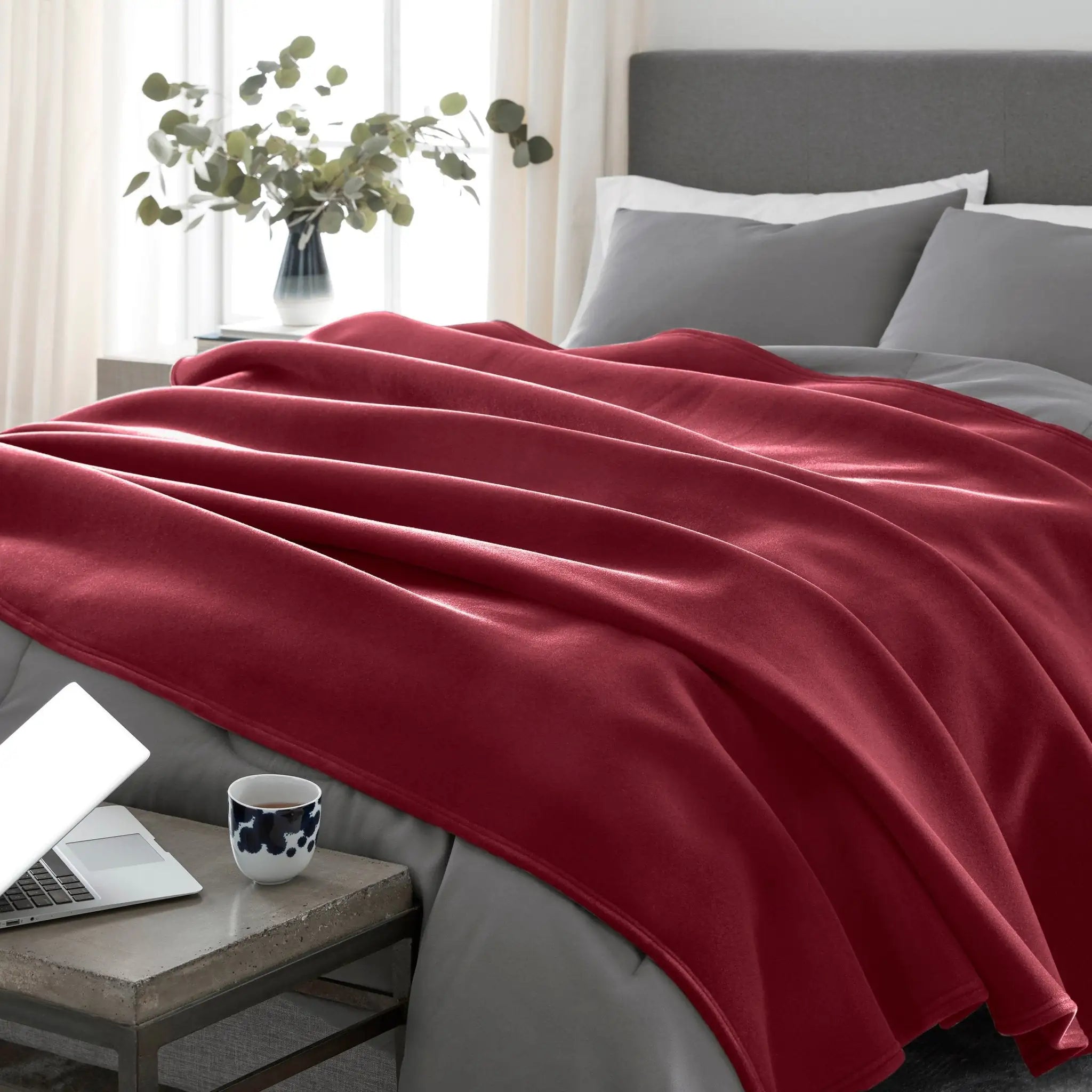 Red blanket draped over a bed with a laptop and mug on a nightstand.