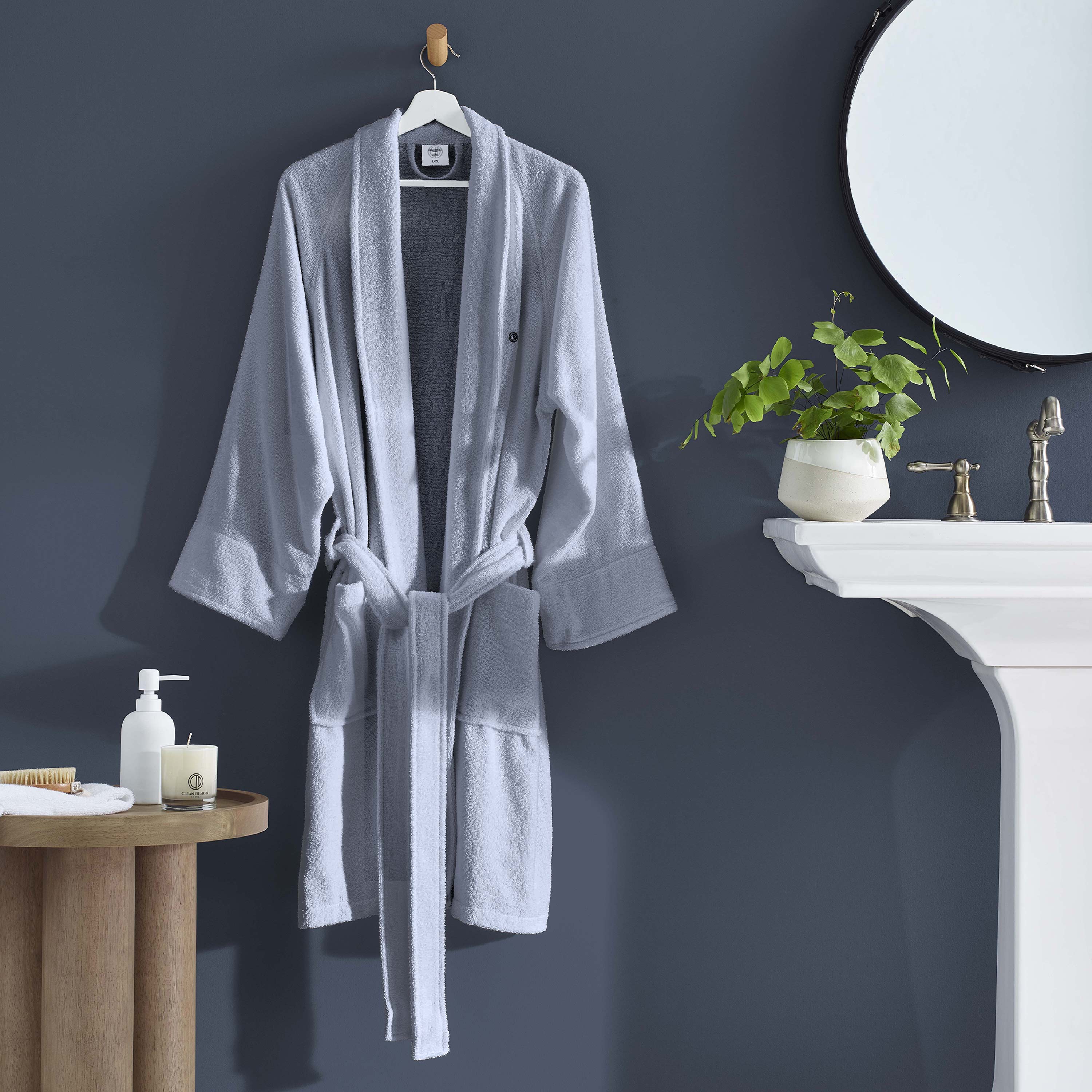 Light gray terry bath robe hanging on a hook against a dark wall, with a sink and plant in the background.