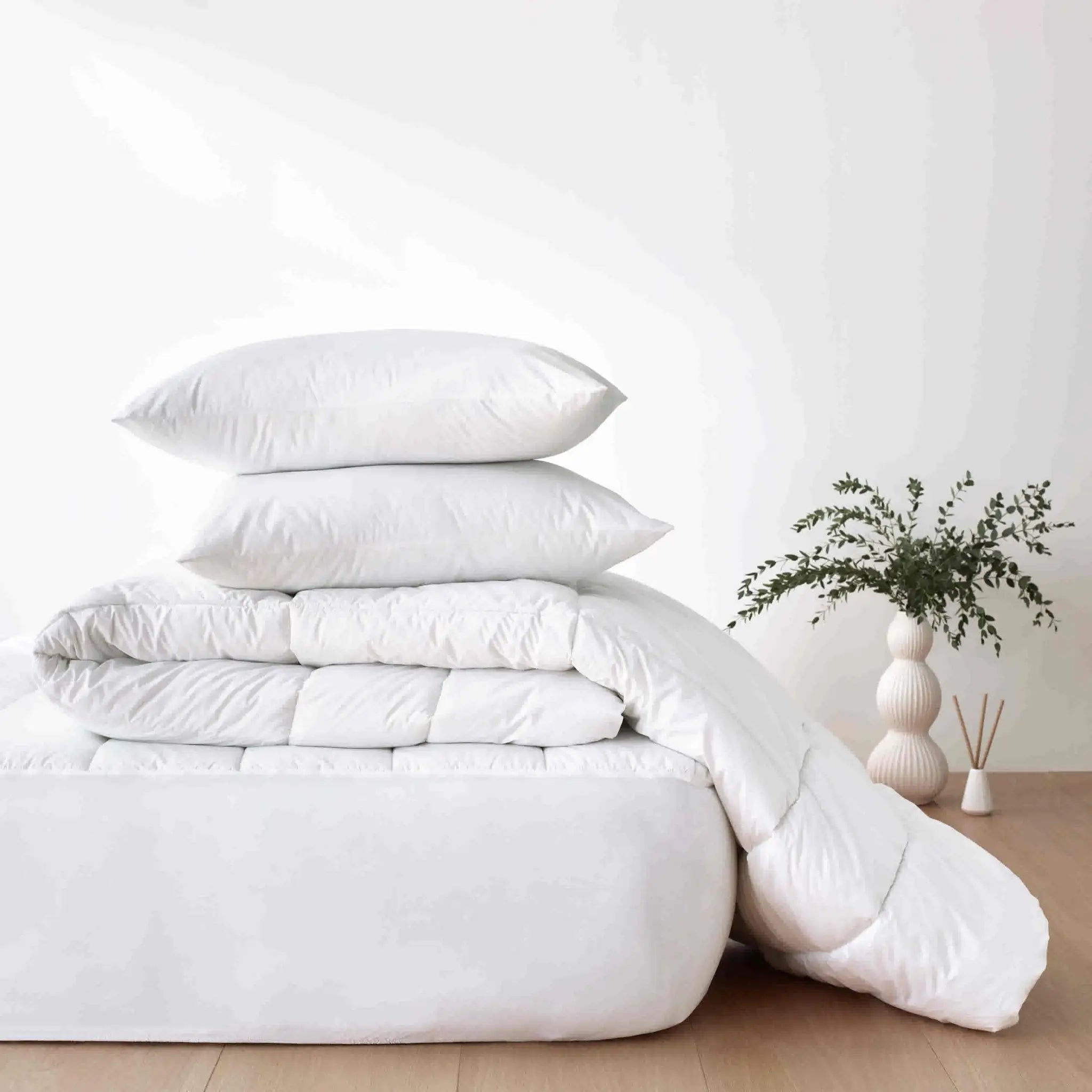A stack of white bedding, including two pillows and a folded quilted comforter, displayed on a mattress in a bright.