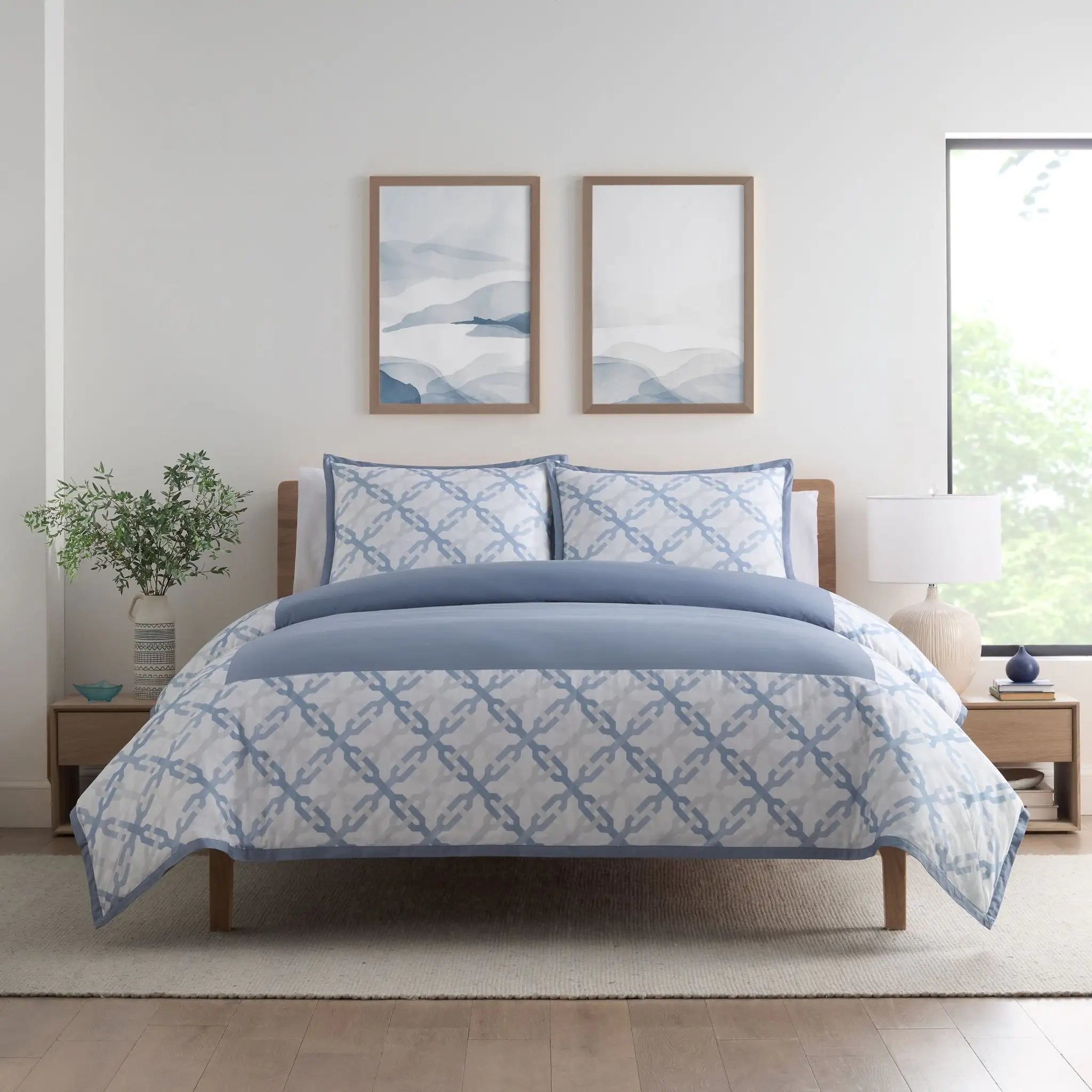A neatly made bed in a bright bedroom, covered with a white comforter and matching pillow shams featuring a light blue geometric trellis pattern and a solid light blue top section.