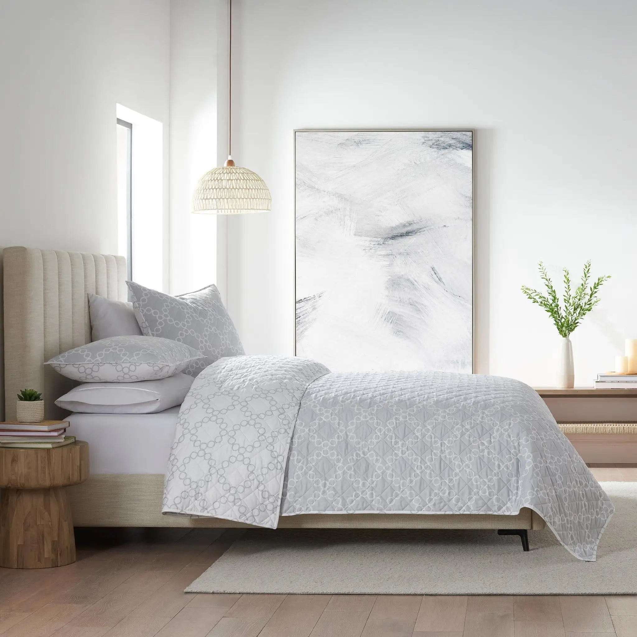 A neatly made bed in a bright, modern bedroom, dressed with a light gray and white patterned quilt and matching pillows.