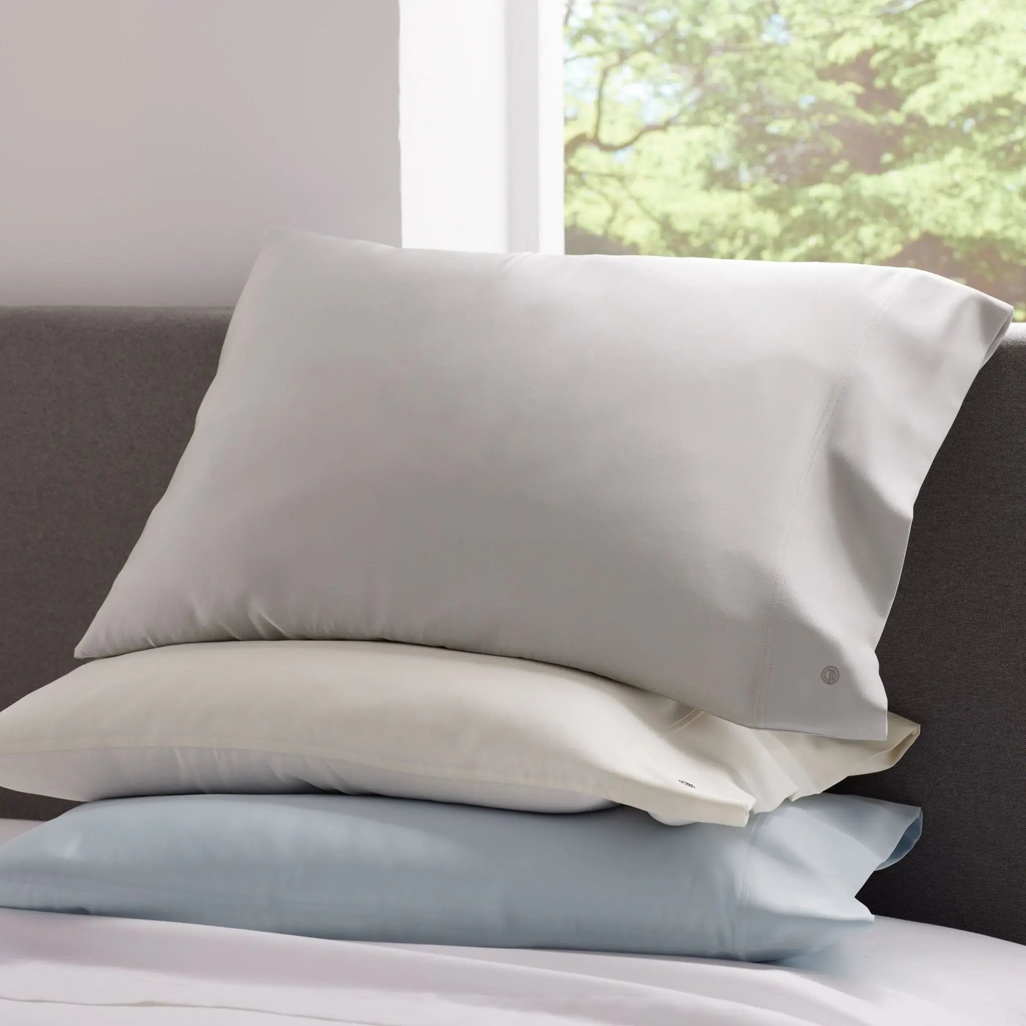 Stack of three pillowcases in gray, white, and light blue on a bed with a window in the background.
