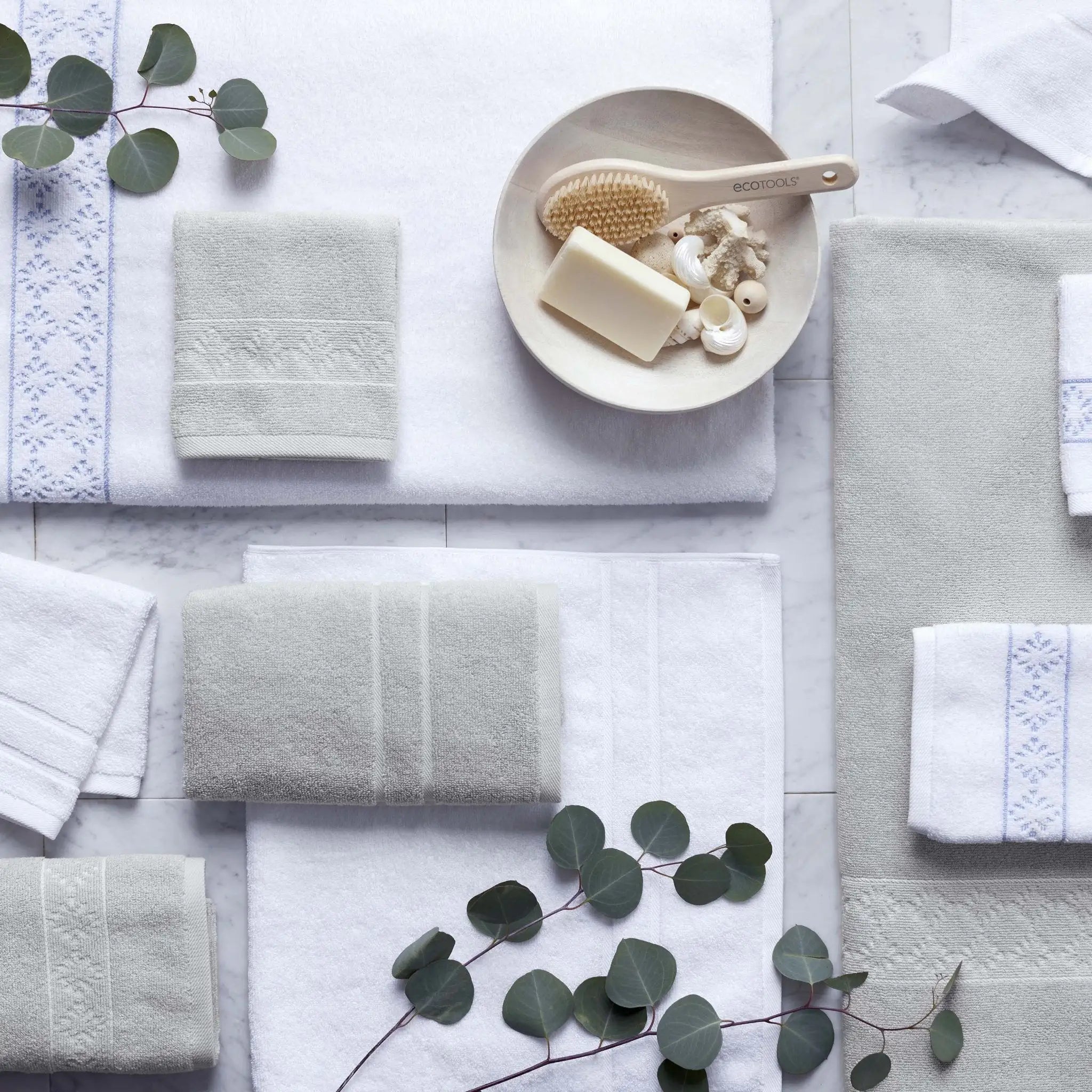 A styled flat-lay of a coordinated bath towel set in soft white and grey, arranged on a light marble surface.