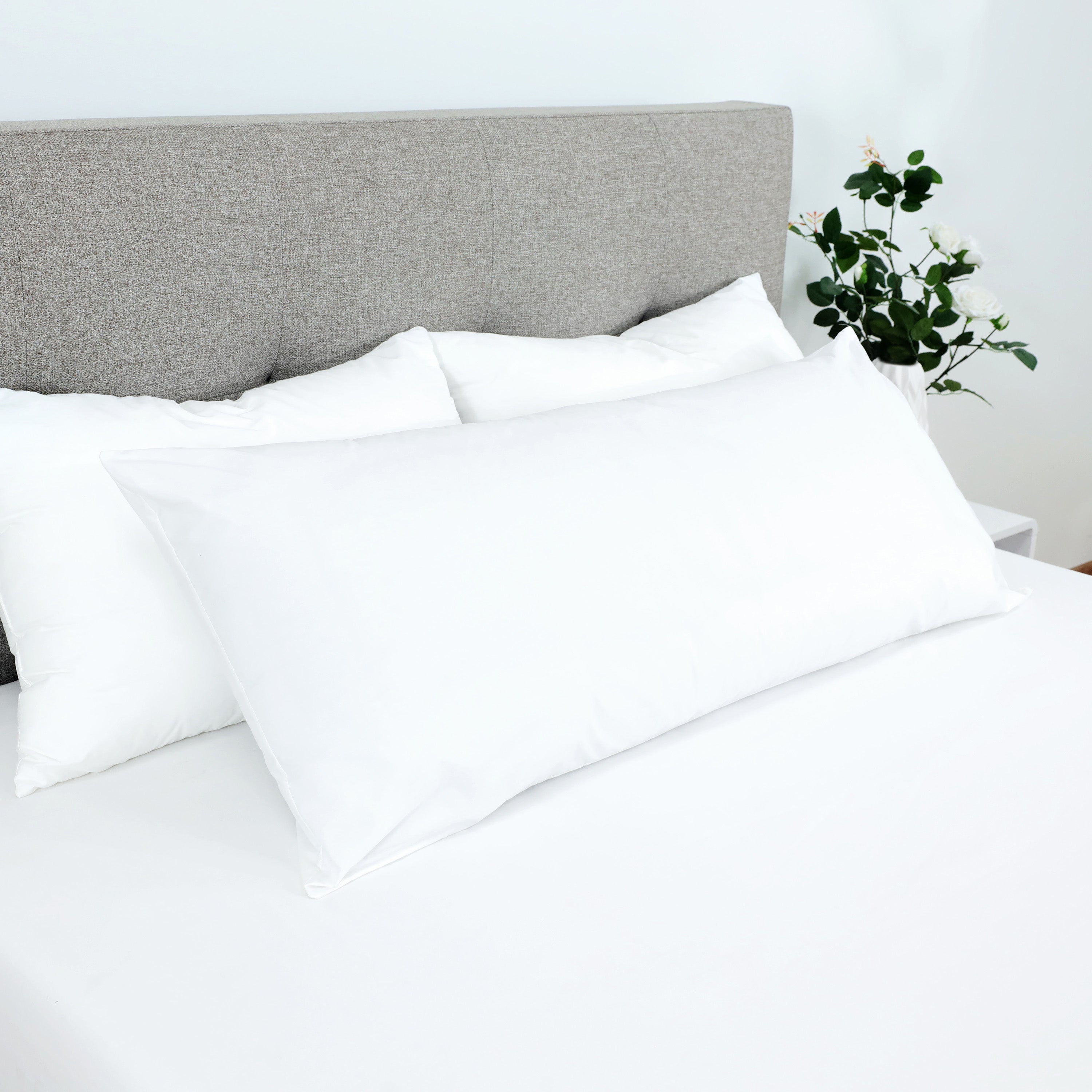 White gusset pillow placed on a bed with a light-colored sheet and a gray upholstered headboard in the background.