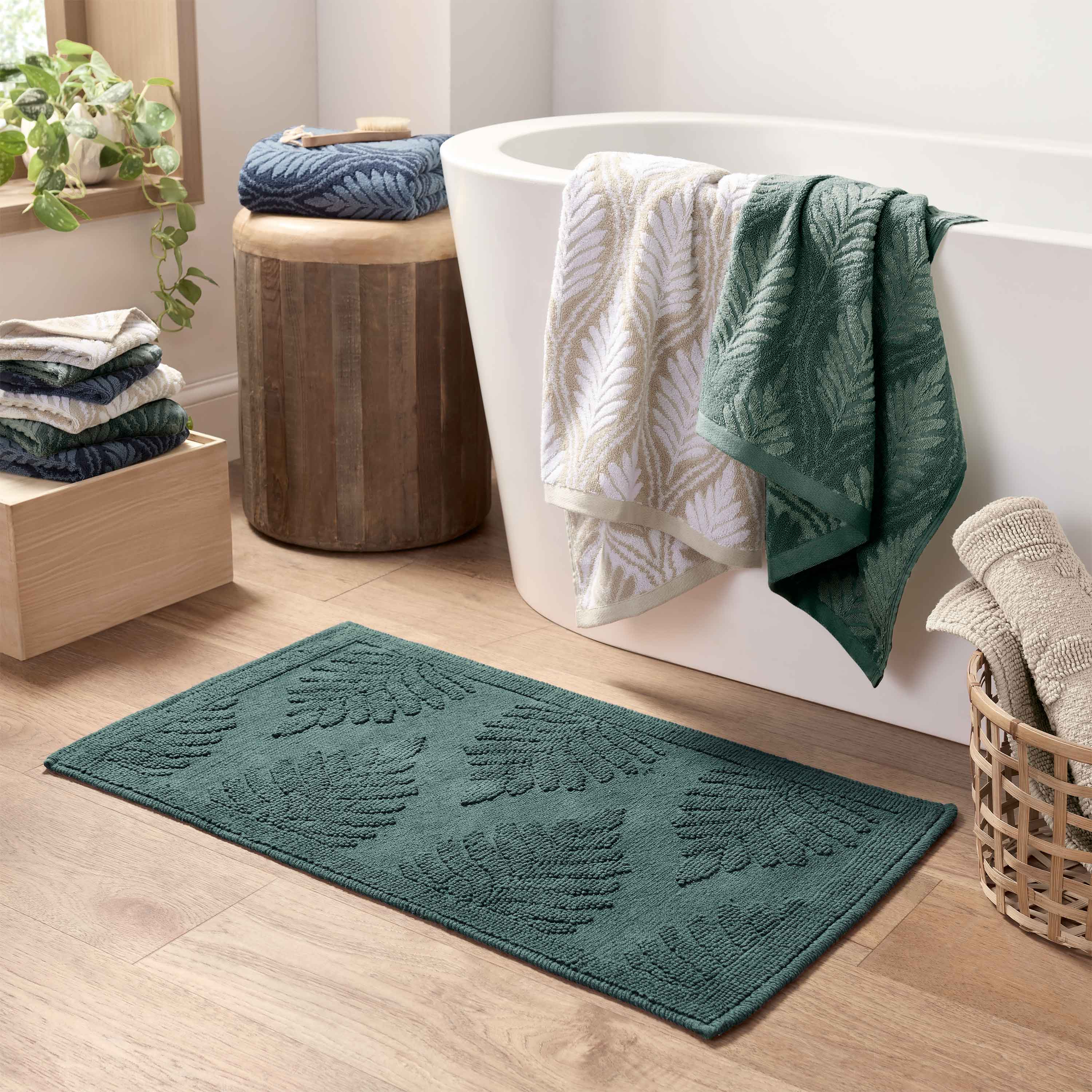 Green bath rug with fern pattern, placed on wooden floor near a bathtub and stacked towels.