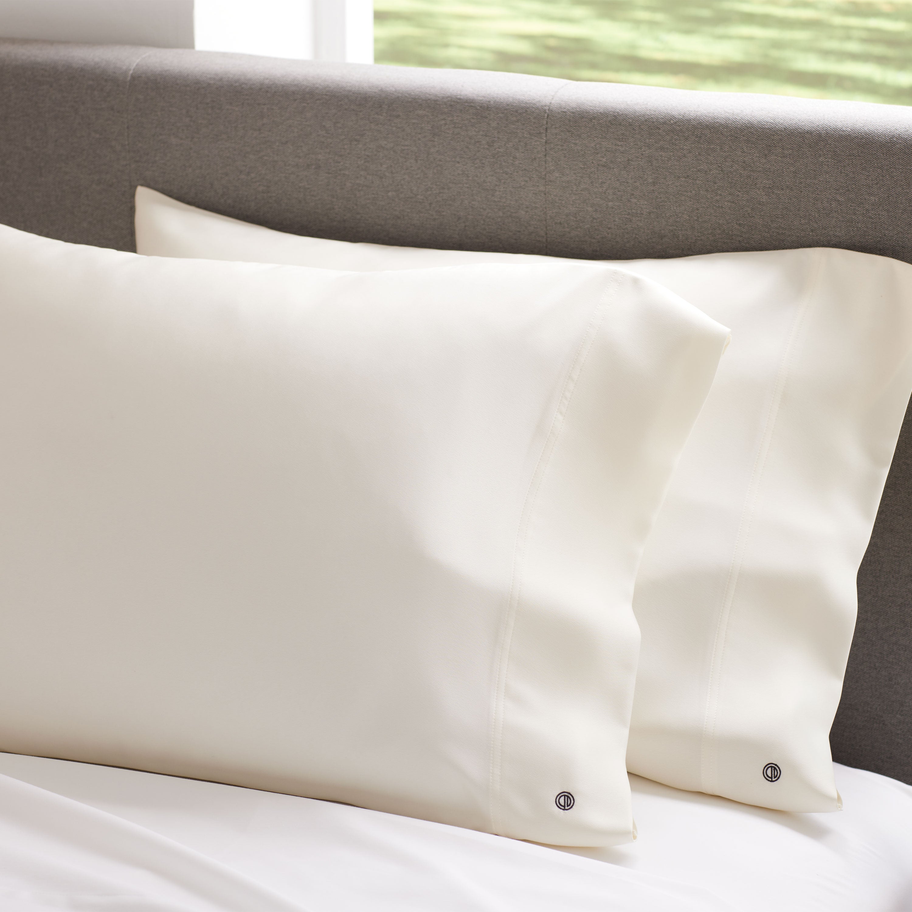 Two cream satin standard pillowcases resting on a white bedspread against a gray headboard.