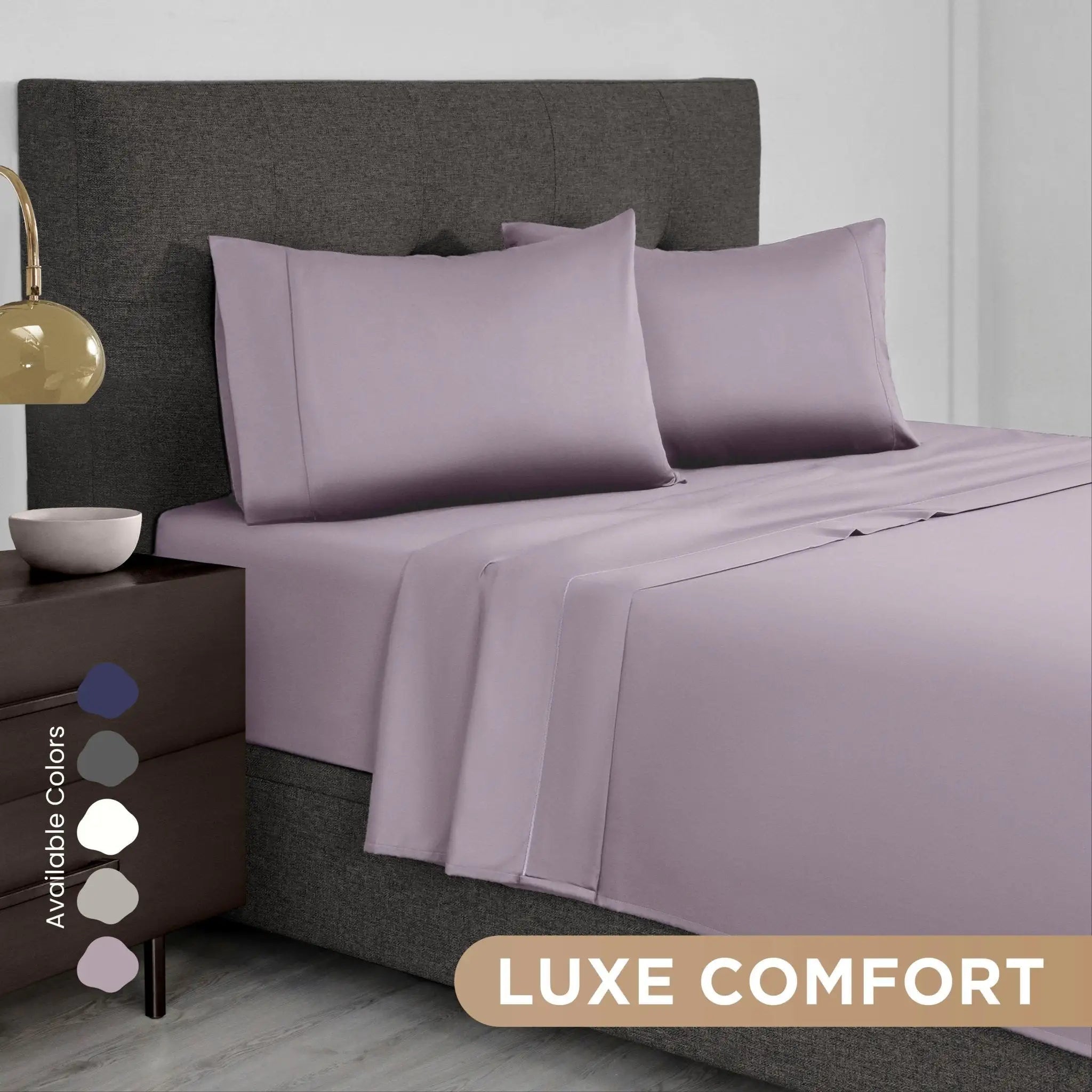 A bed dressed in a lilac bedding set with two pillowcases and a matching sheet, shown in a styled bedroom scene with a dark upholstered headboard, bedside lamp, and a text badge reading 'LUXE COMFORT' plus an 'Available Colors' swatch panel.