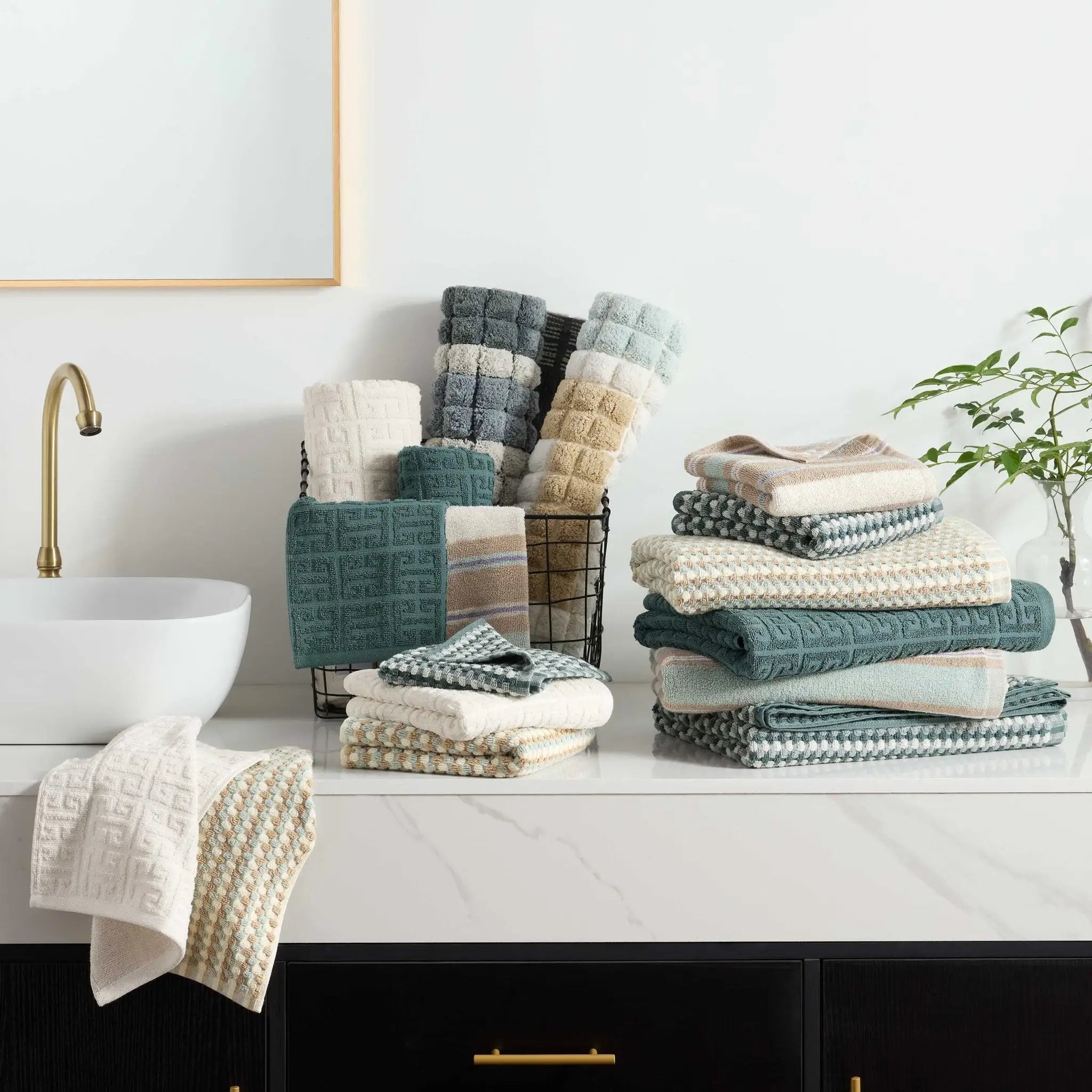 A collection of textured bath towels in neutral and muted teal tones, neatly stacked on a white marble bathroom counter.