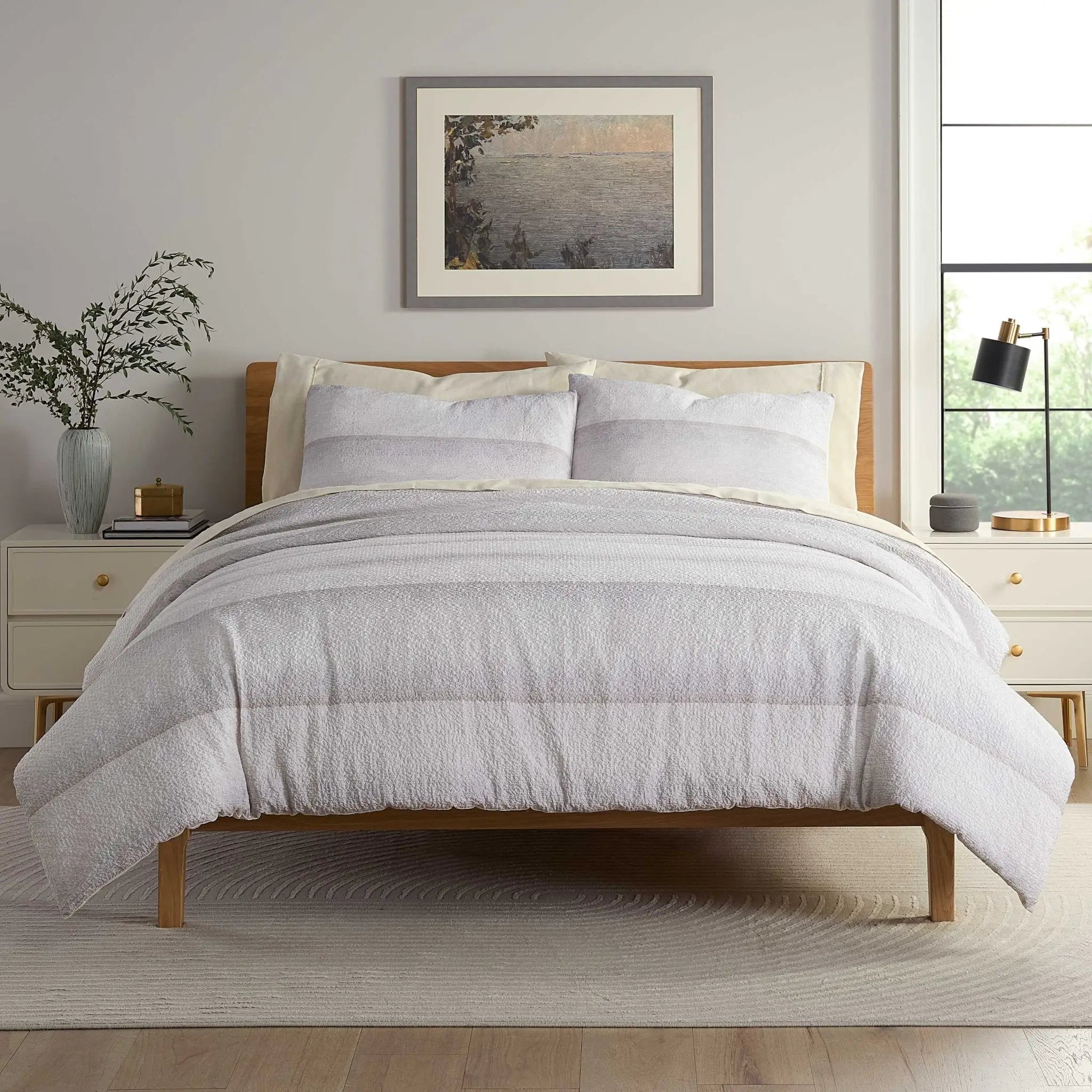 A neatly made bed with a light gray textured comforter and matching pillow shams, set in a bright bedroom.