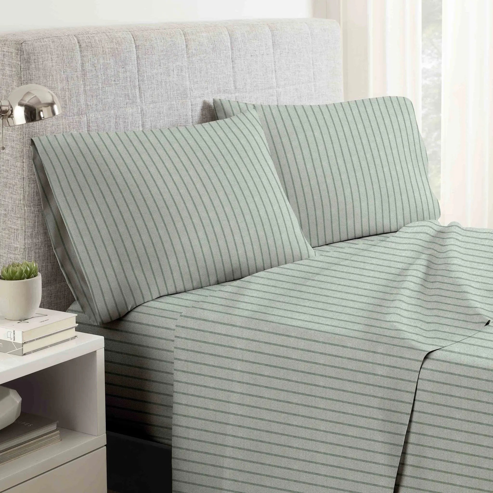 A bed dressed in a light sage green sheet set with thin vertical dark green stripes, styled with two matching pillowcases in front of a gray upholstered headboard.