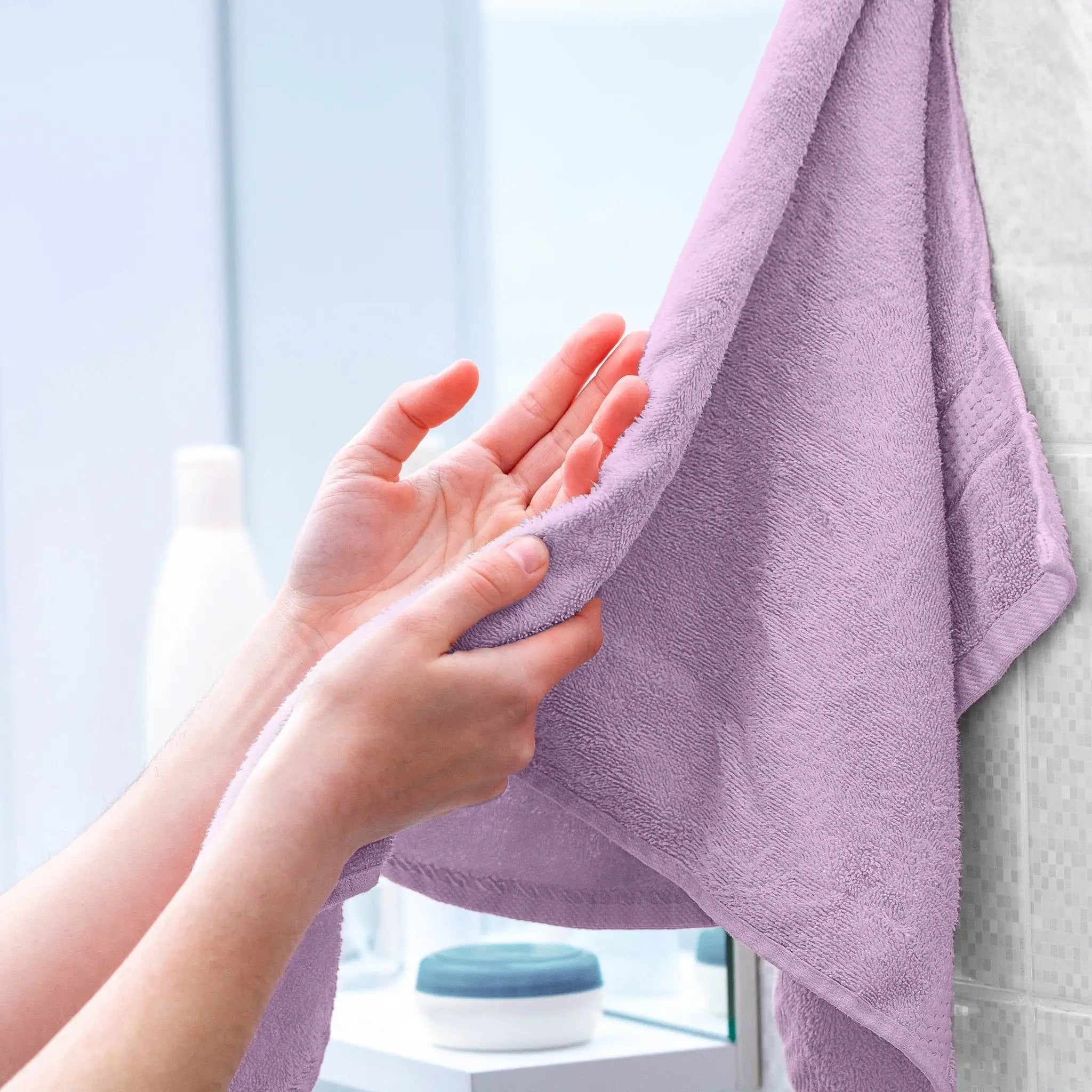 A close-up of a person's hands holding and smoothing a lilac towel, with a blurred bathroom background.
