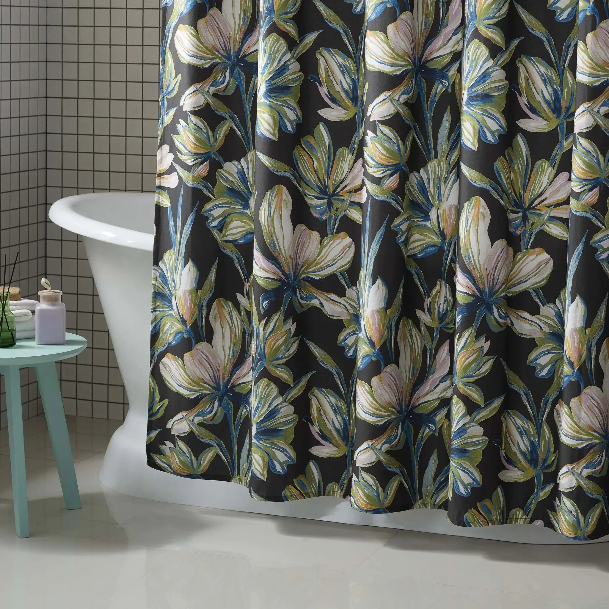 A floral shower curtain hanging in a bathroom, featuring large white and blue flowers with yellow-green centers on a dark charcoal background. A white clawfoot tub, a small mint green stool, and tiled walls are visible.