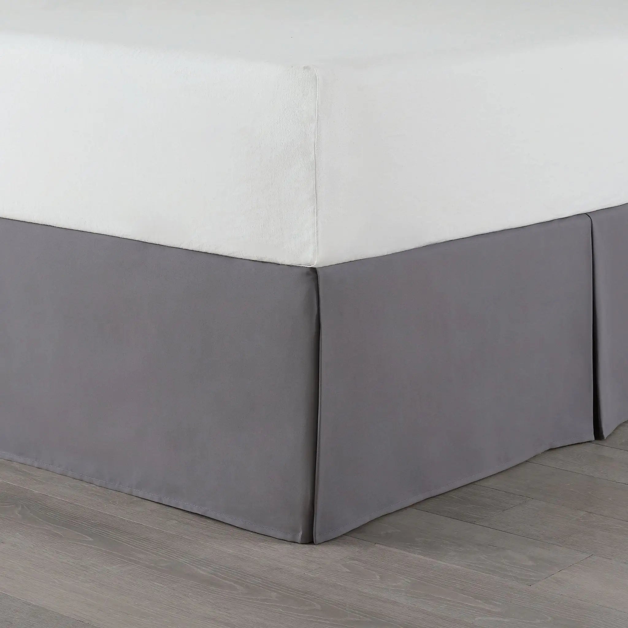 A gray bed skirt made of fabric, shown on a bed with a white fitted sheet and gray wood-look flooring.