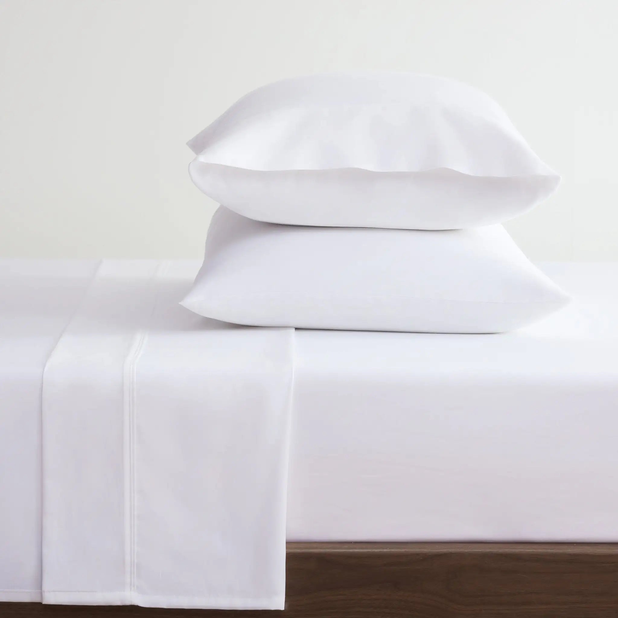 Two white pillows stacked on a white bed sheet with a folded white pillowcase, set against a plain light background.