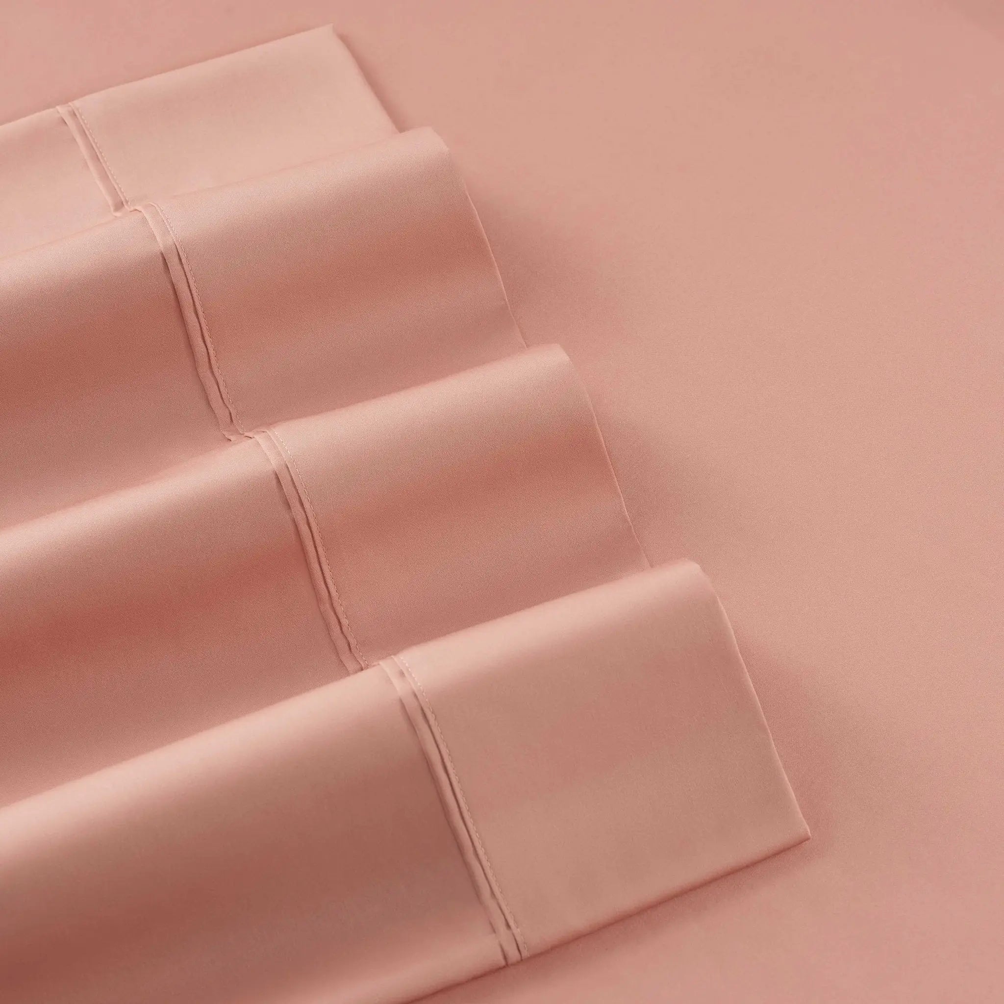 A close-up of folded dusty pink bedding, likely a pillowcase or sheet set, arranged diagonally on a matching solid pink background.