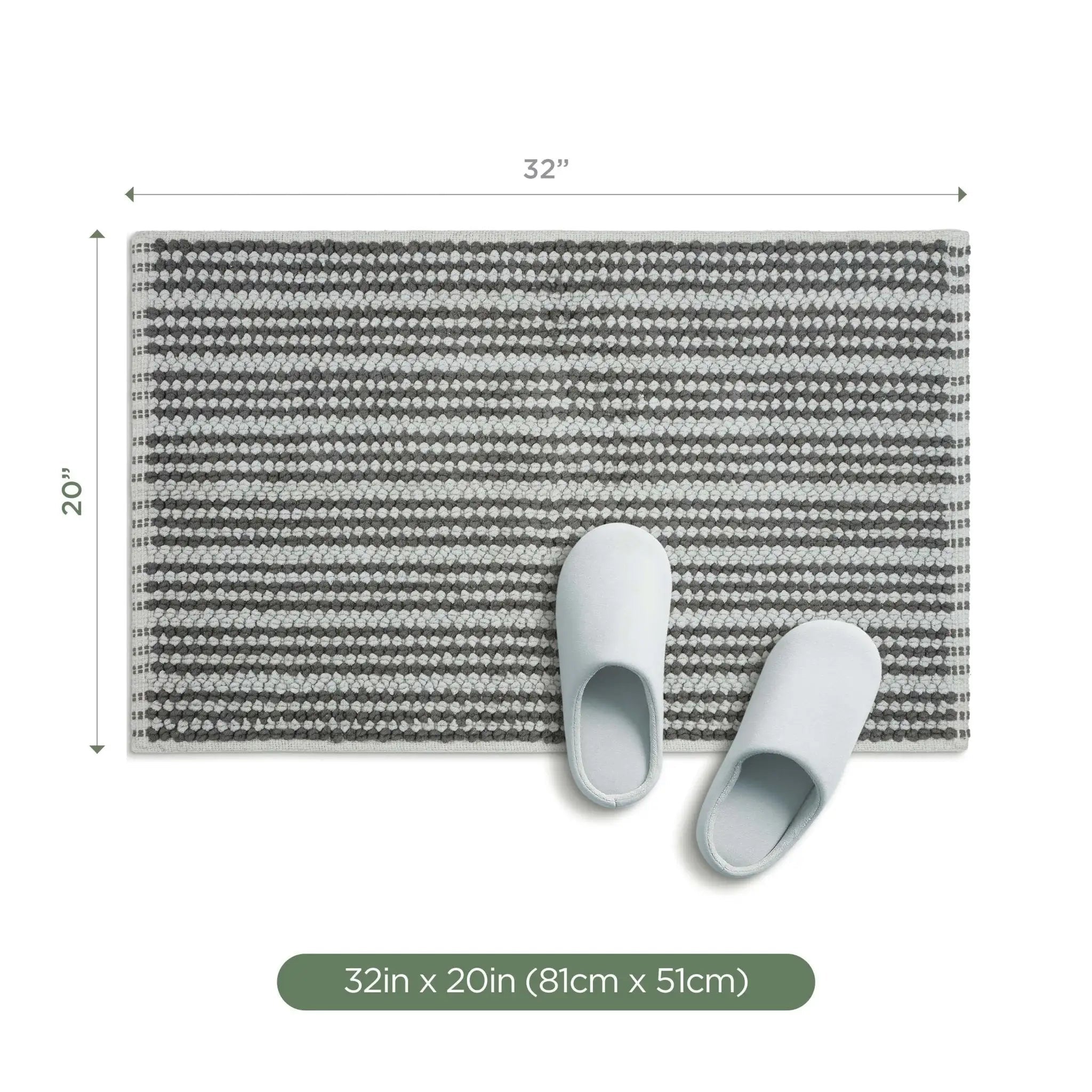 A rectangular bath rug with a charcoal striped pattern, shown with dimension markers of 32 inches by 20 inches, with a pair of light gray slip-on slippers placed on top. A green label at the bottom reads '32in x 20in (81cm x 51cm)'.