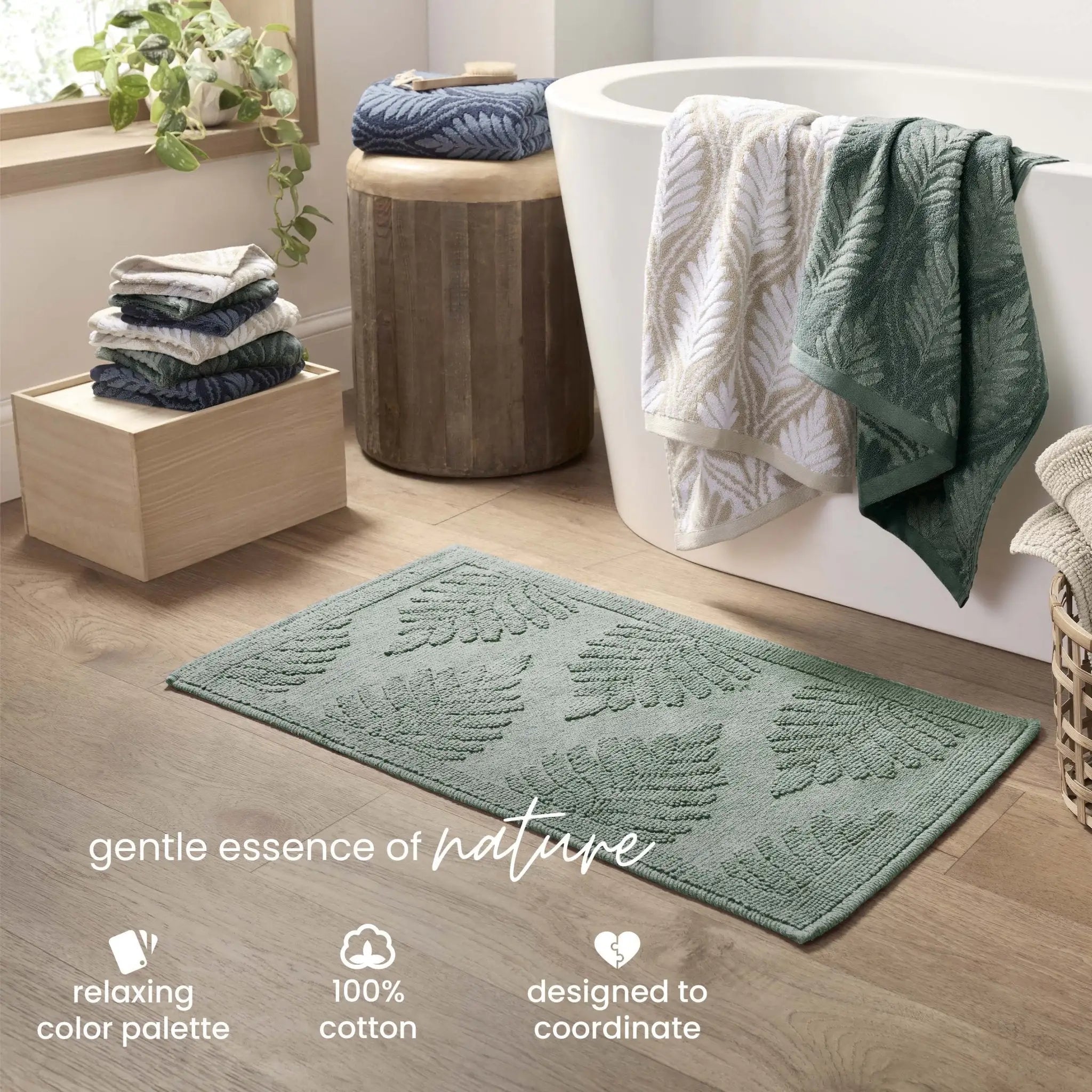 A sage bath rug on a light wood floor in a bathroom setting, with a white freestanding tub, folded towels, and decorative text at the bottom.