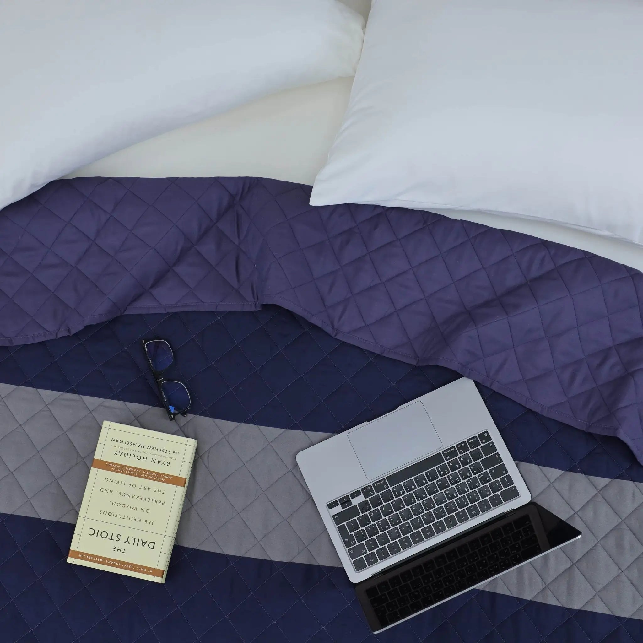 A styled top-down view of a bed with a quilted coverlet in horizontal stripes of white, deep purple, and navy blue.