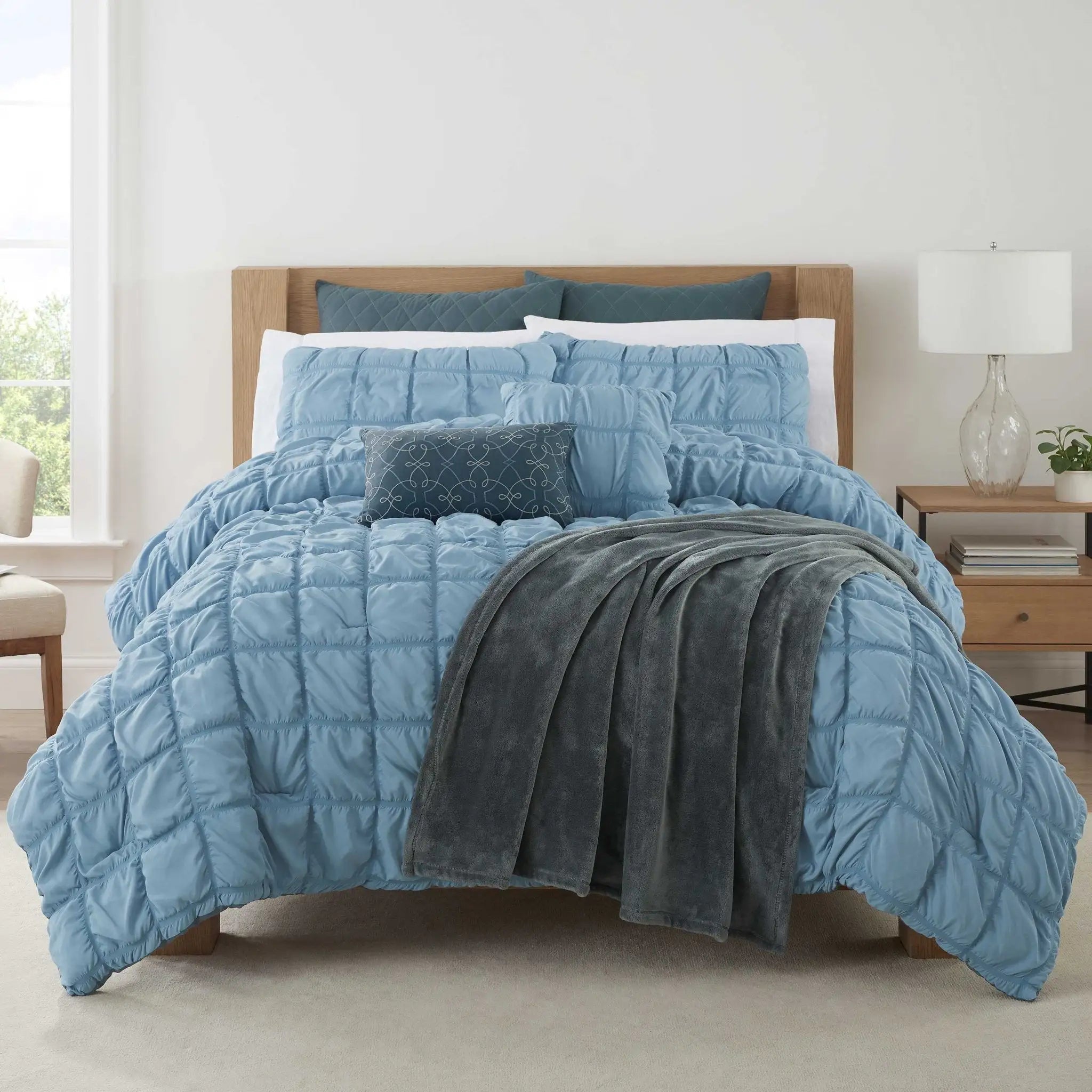 A bed dressed in a light blue quilted comforter set with matching pillows, a dark blue patterned accent pillow, and a dark gray throw blanket, shown in a bright bedroom setting.