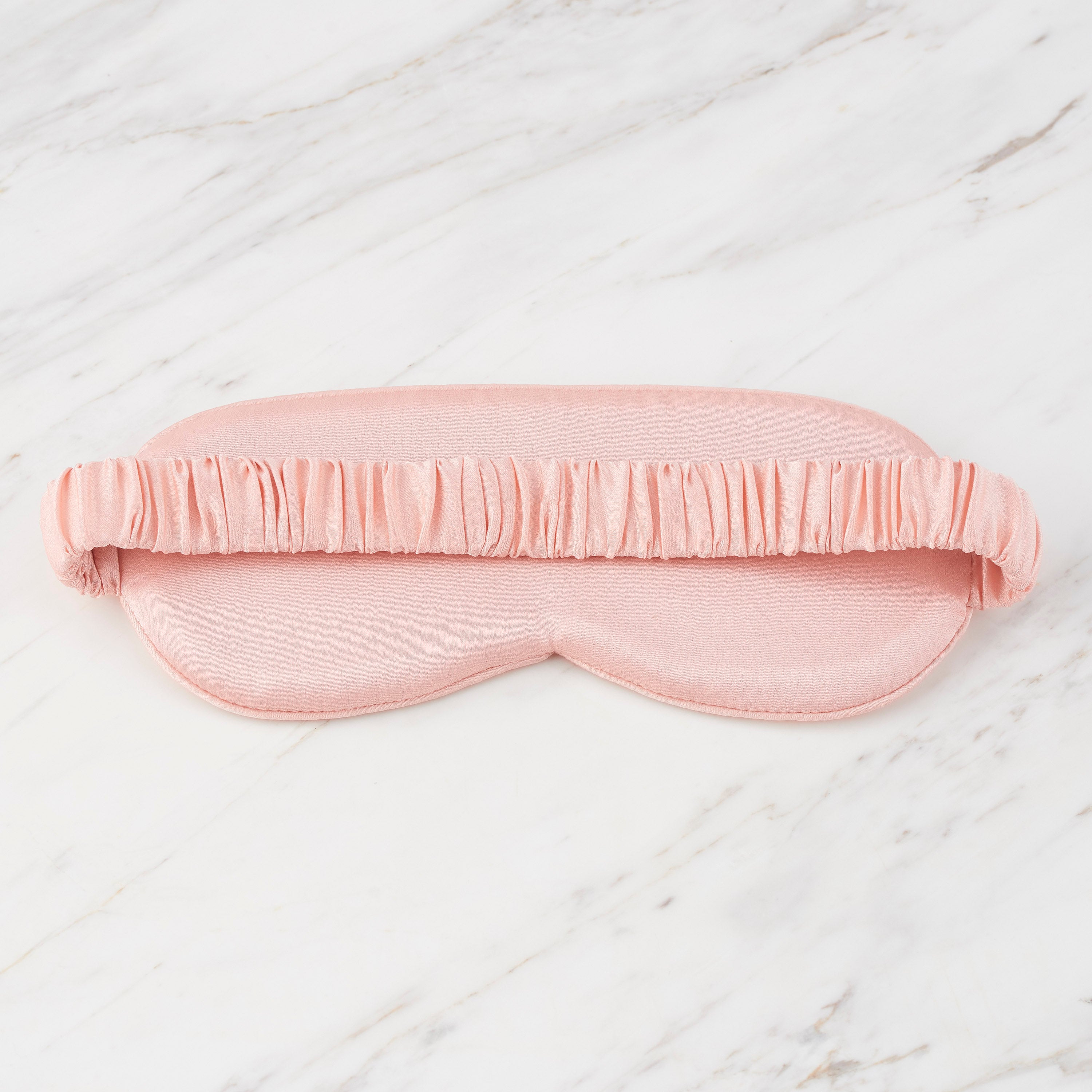 Pink satin eye mask with a ruffled elastic band, resting on a marble surface.