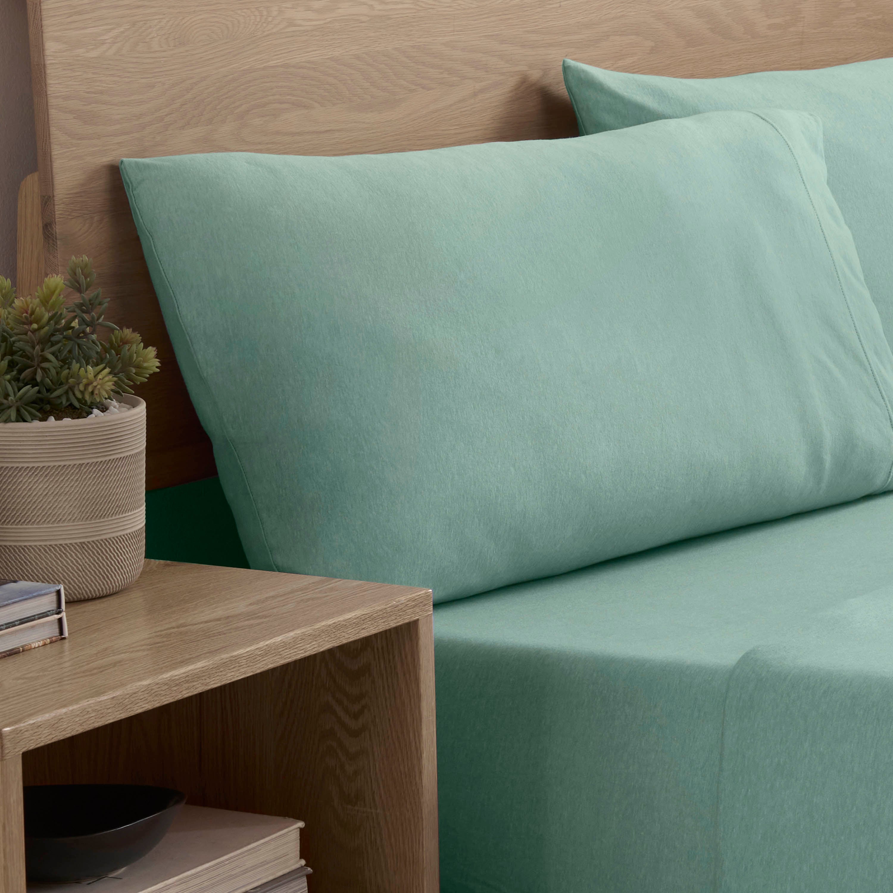 Light green sheet set with two matching pillows on a bed, next to a wooden nightstand with a plant and books.