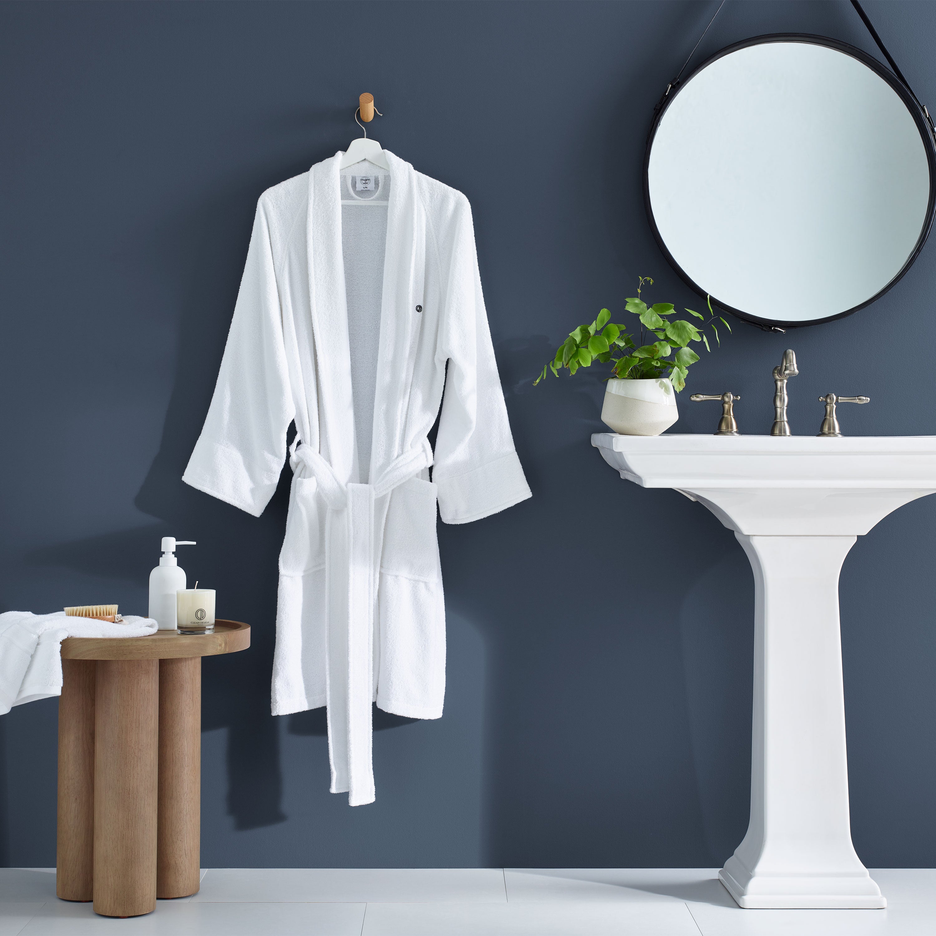 White terry bath robe hanging on a hook, with a round mirror, sink, and plant in a modern bathroom setting.