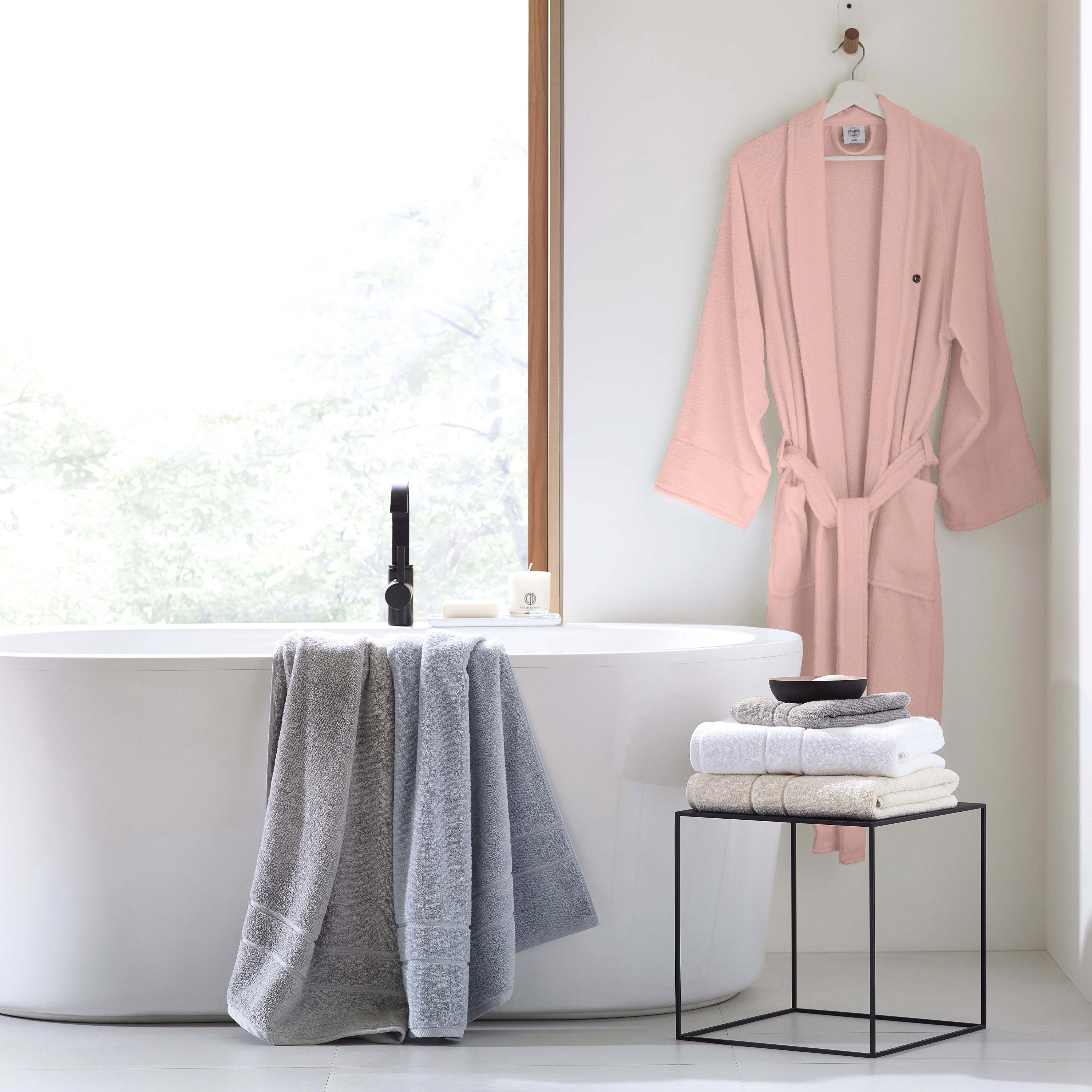 Light pink terry bath robe hangs on a hook next to a freestanding tub with stacked towels on a black metal stand.