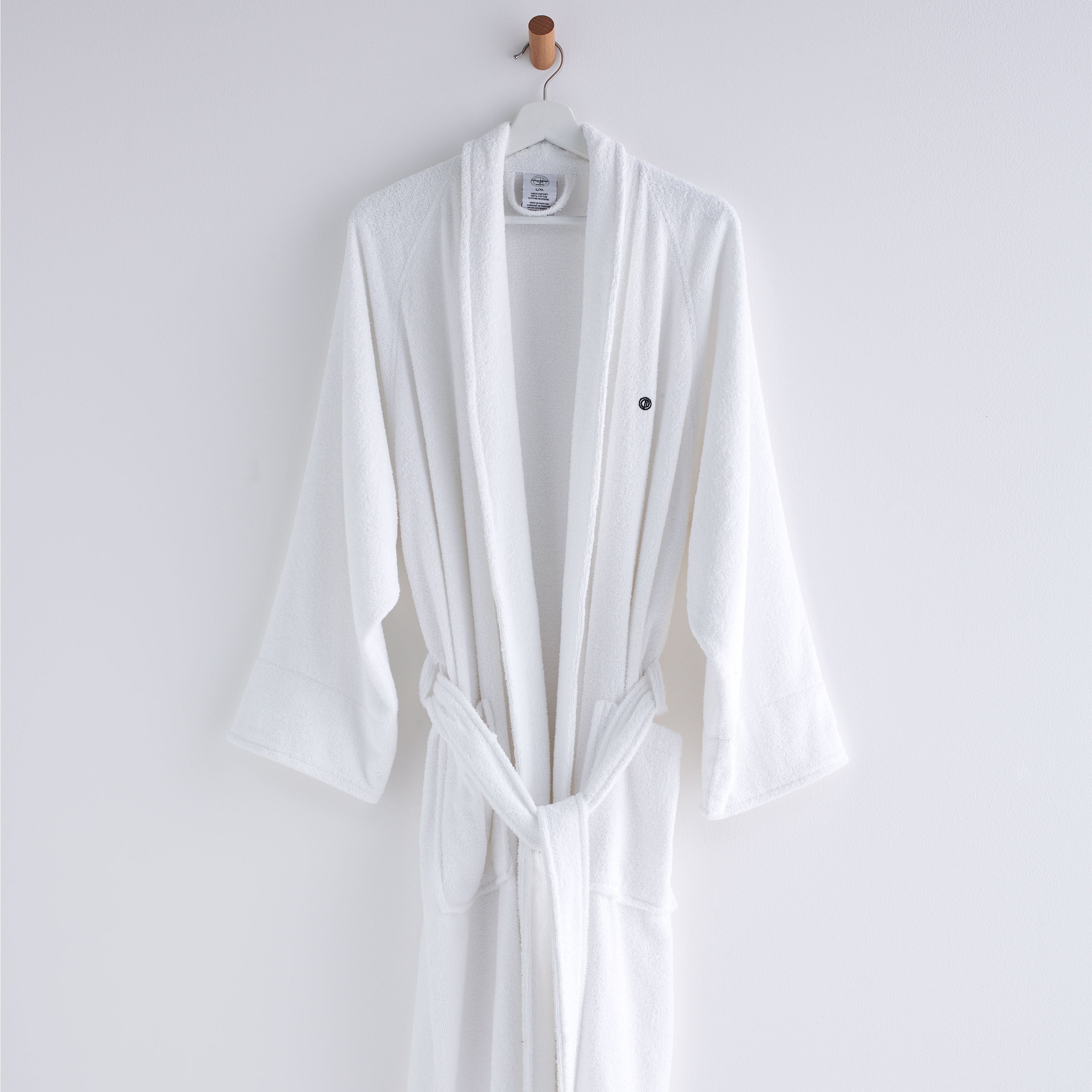 White terry bath robe with long sleeves and a tied waist, hanging on a hook against a plain background.